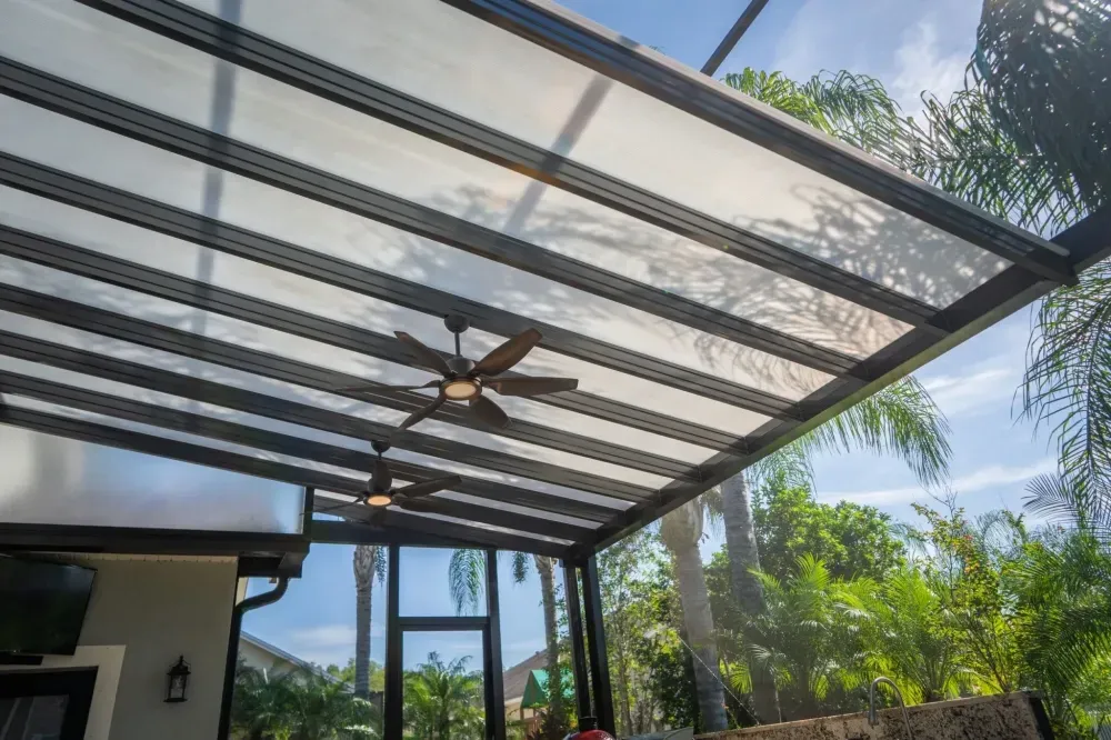 Covered patio with clear roof panels, ceiling fans, and lush greenery in bright daylight