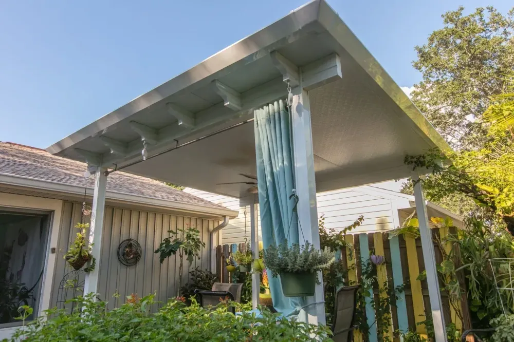 Covered patio with translucent blue curtains, surrounded by greenery and a house exterior.
