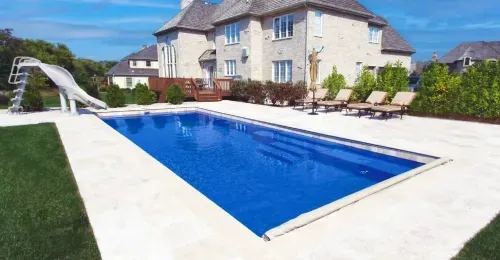 Backyard with a blue rectangular swimming pool beside a large stone house and patio