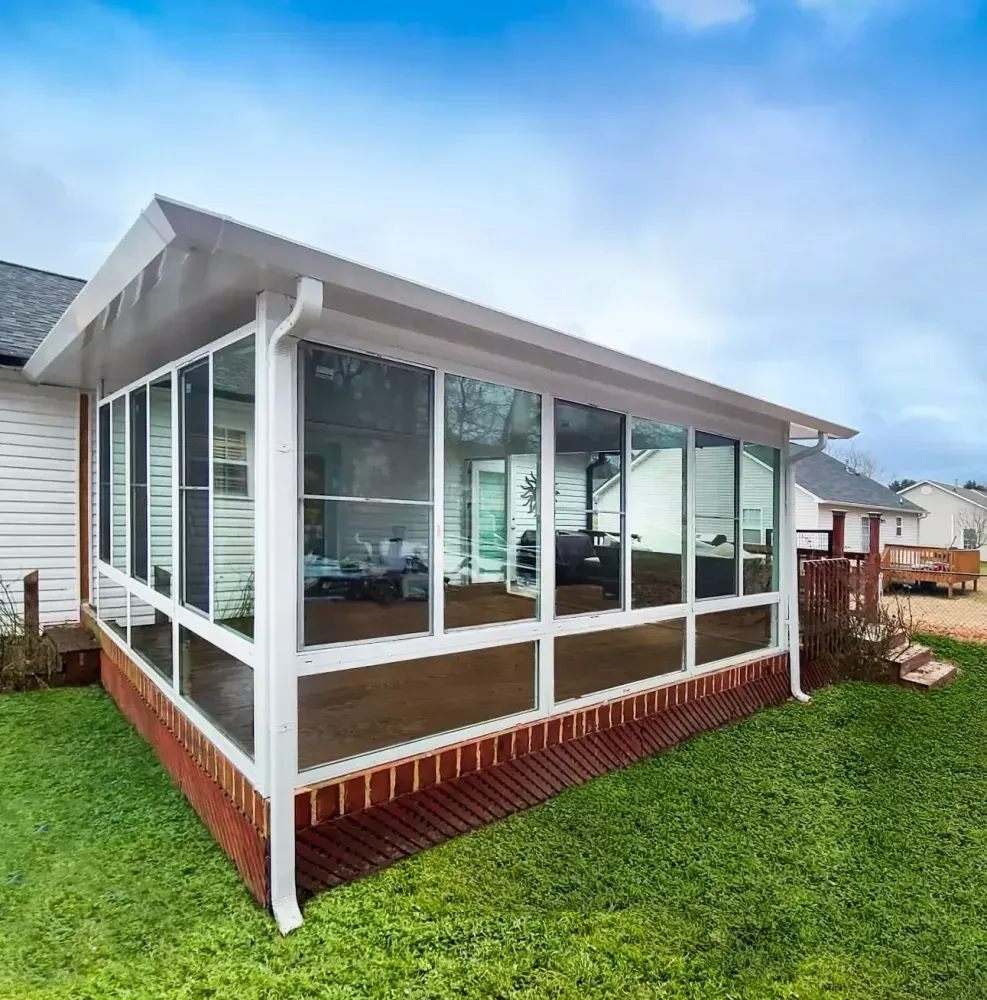Glass-enclosed sunroom with white trim and brick skirting beside a grassy yard