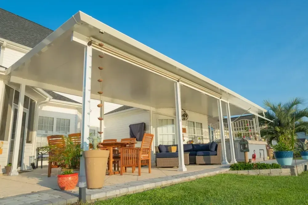 Covered patio with wooden dining set, outdoor sofa, and potted plants beside a house under a blue sky