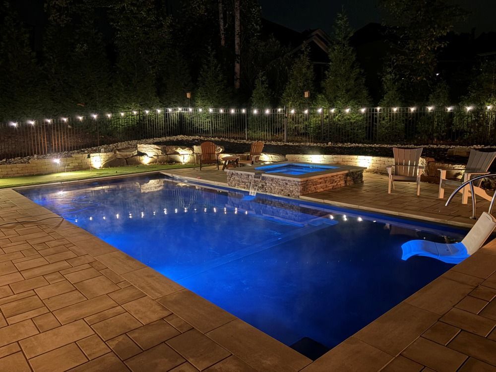 Illuminated backyard pool at night with blue water, patio seating, and string lights along the fence