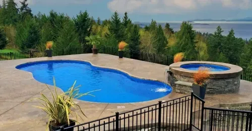 Backyard pool with curved blue water, round hot tub, and stone patio overlooking trees