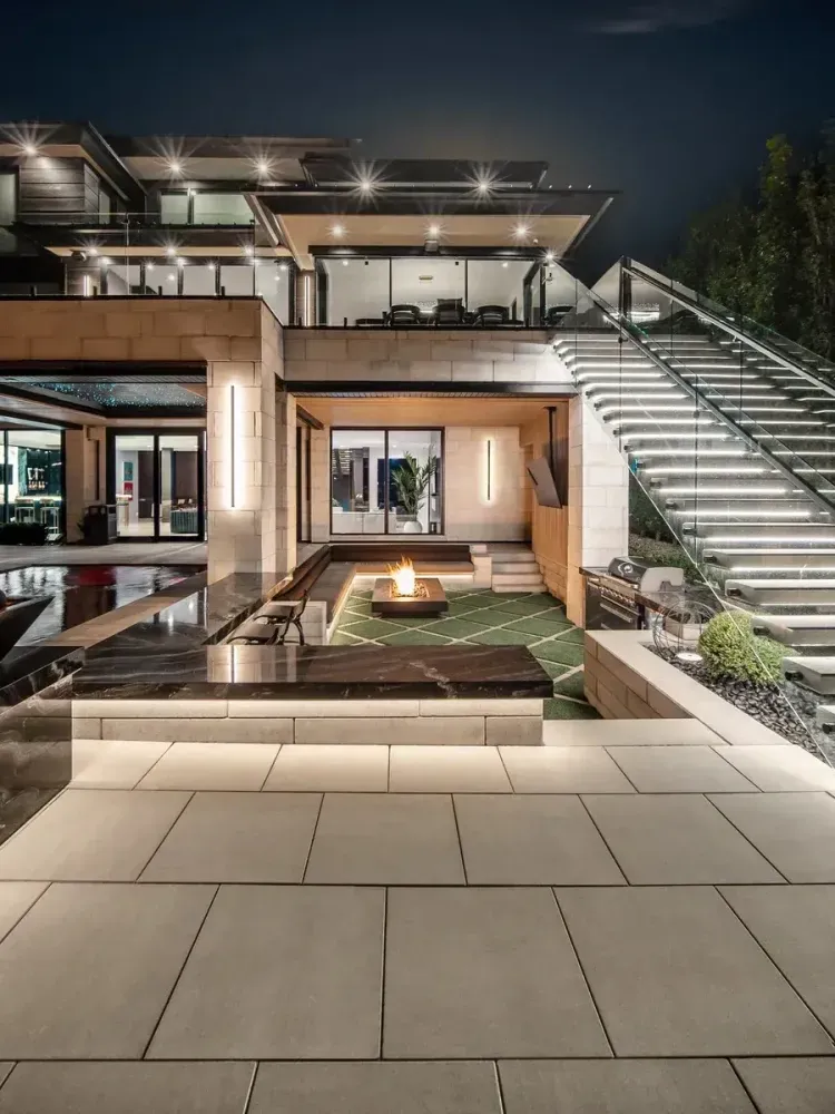 Modern luxury home patio at night with an outdoor staircase, lit seating area, and large stone tiles