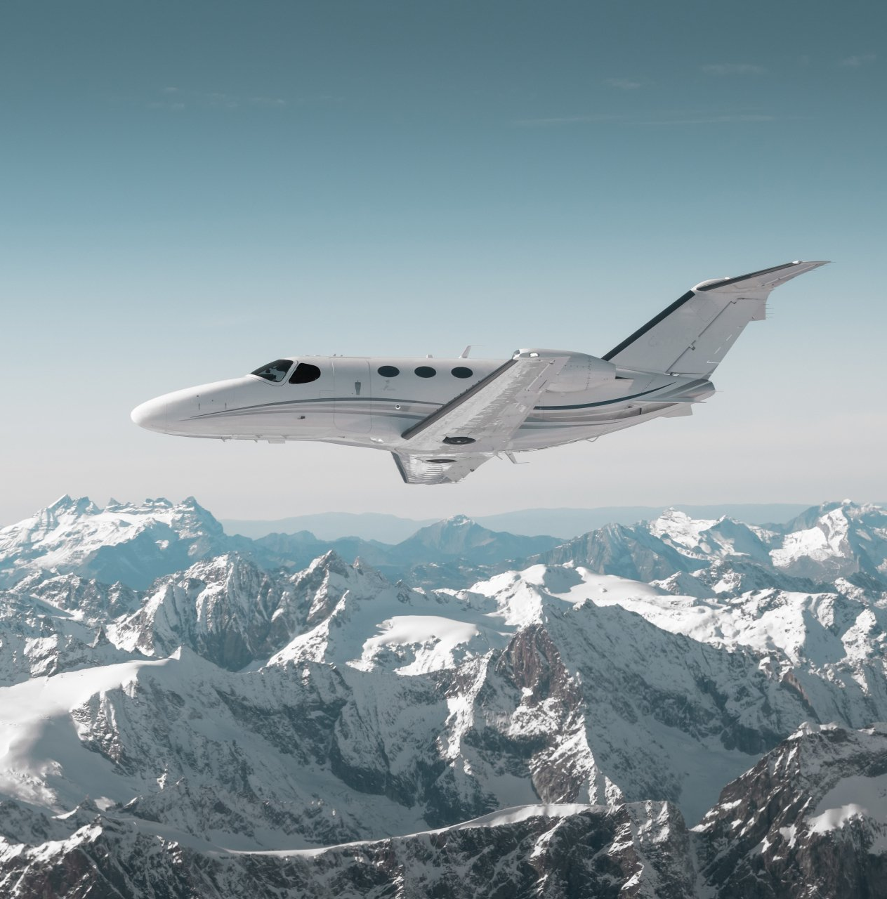 A white private jet flying above snowy mountain peaks under a clear sky
