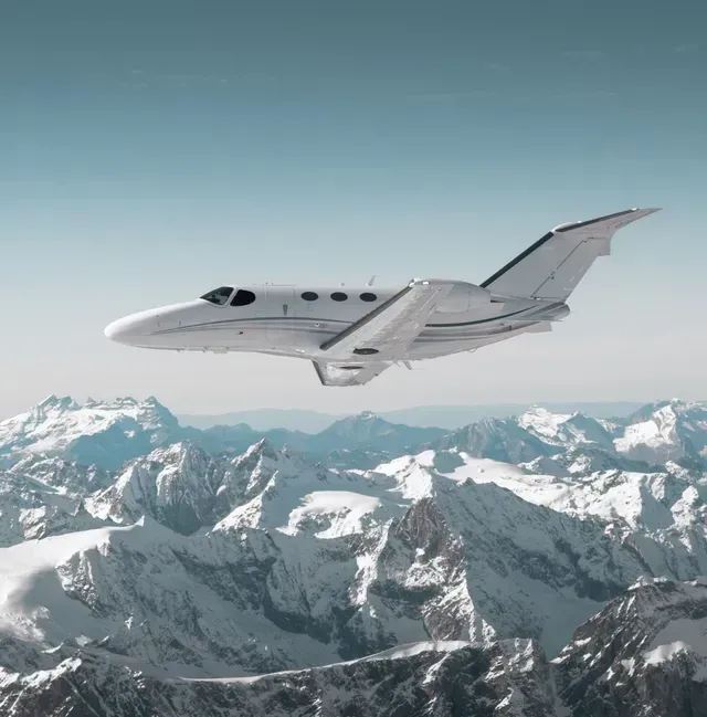 A white private jet flying above snowy mountain peaks under a clear sky