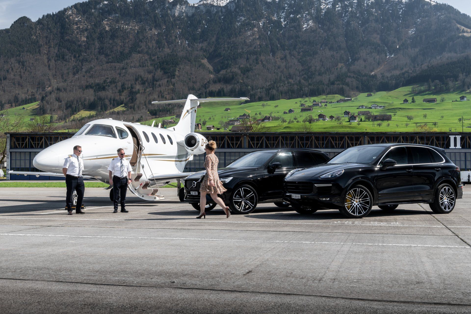 Private jet on tarmac with two black SUVs and people boarding, mountains in the background