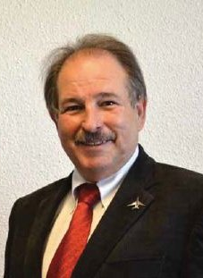 Smiling man in a dark suit and red tie against a plain light wall