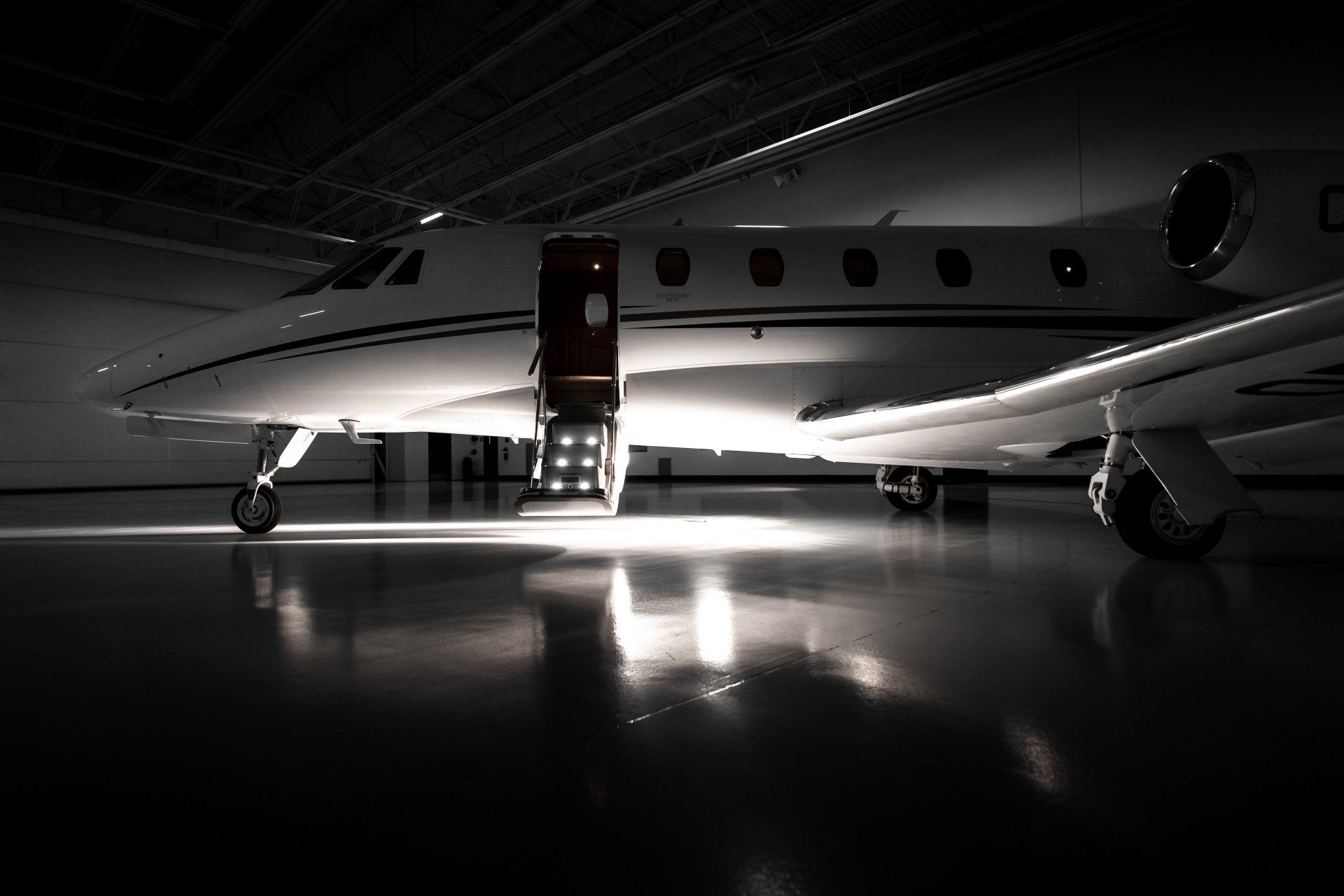 Private jet in a dim hangar with open door and staircase, reflected on polished floor