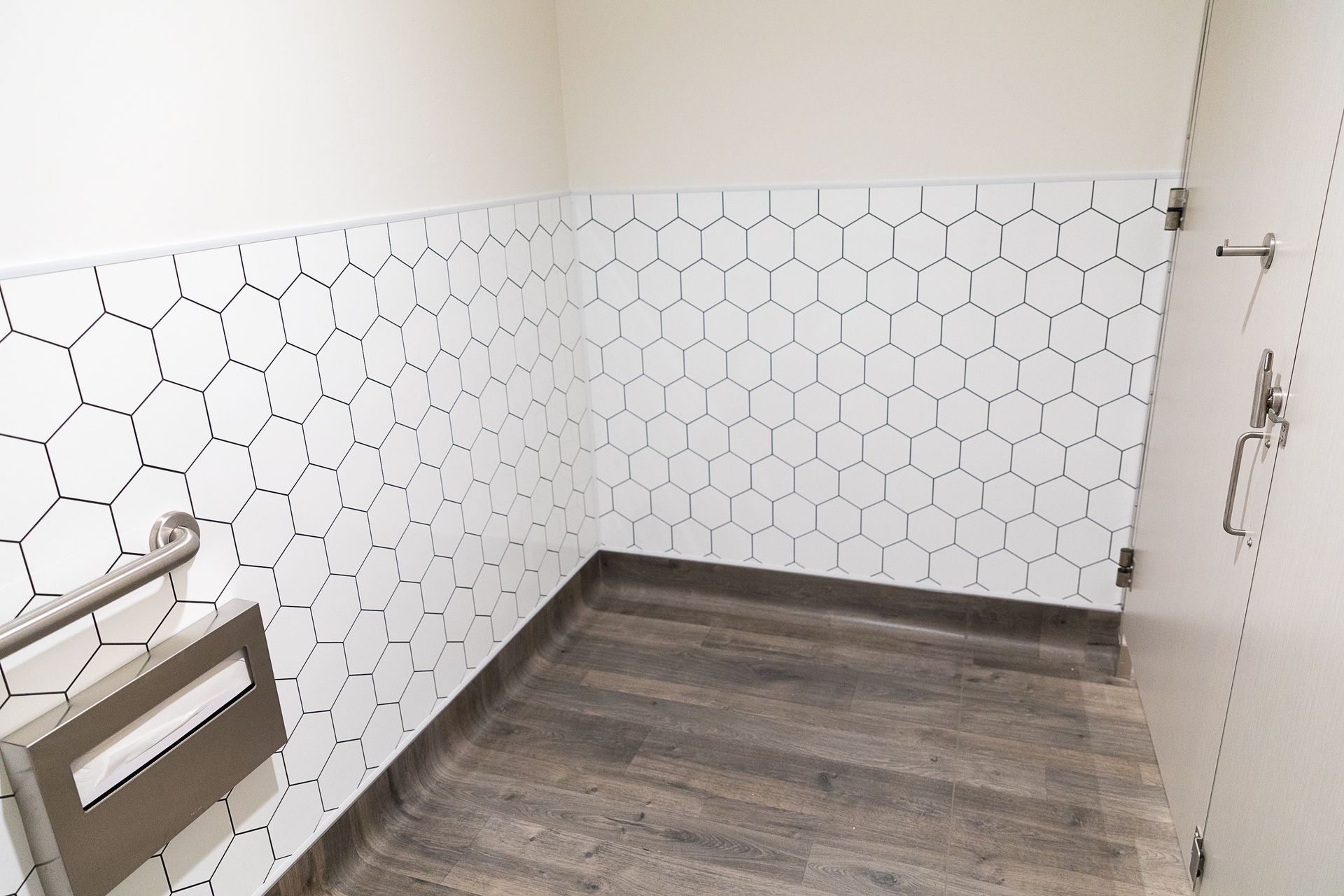 Empty bathroom with white hex tile walls, wood-look floor, and a grab bar near the toilet area