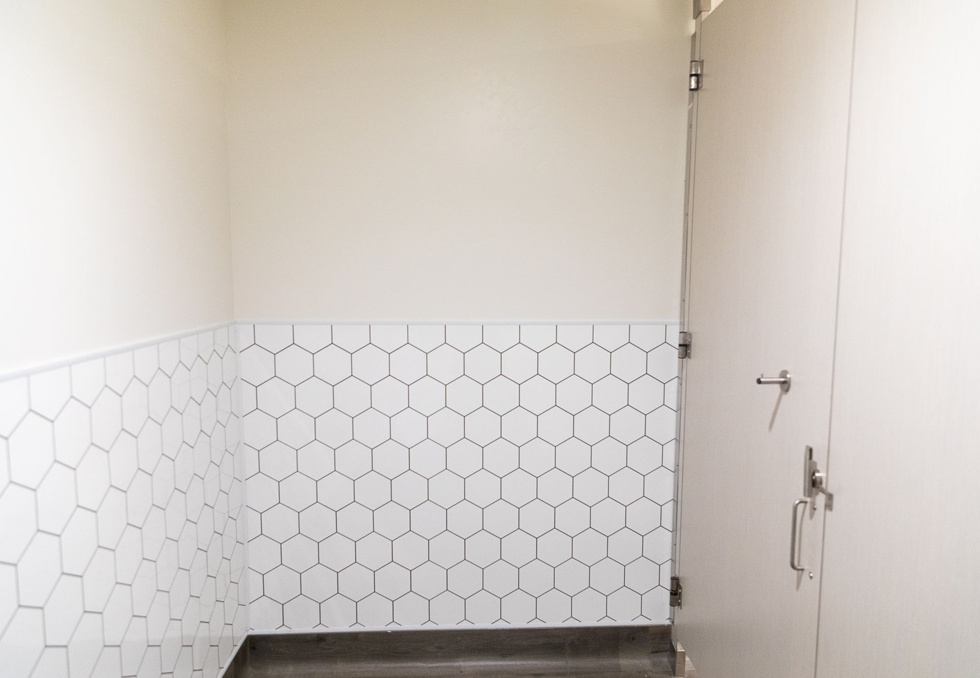 Small white tiled room with a door and metal hook on the right wall.