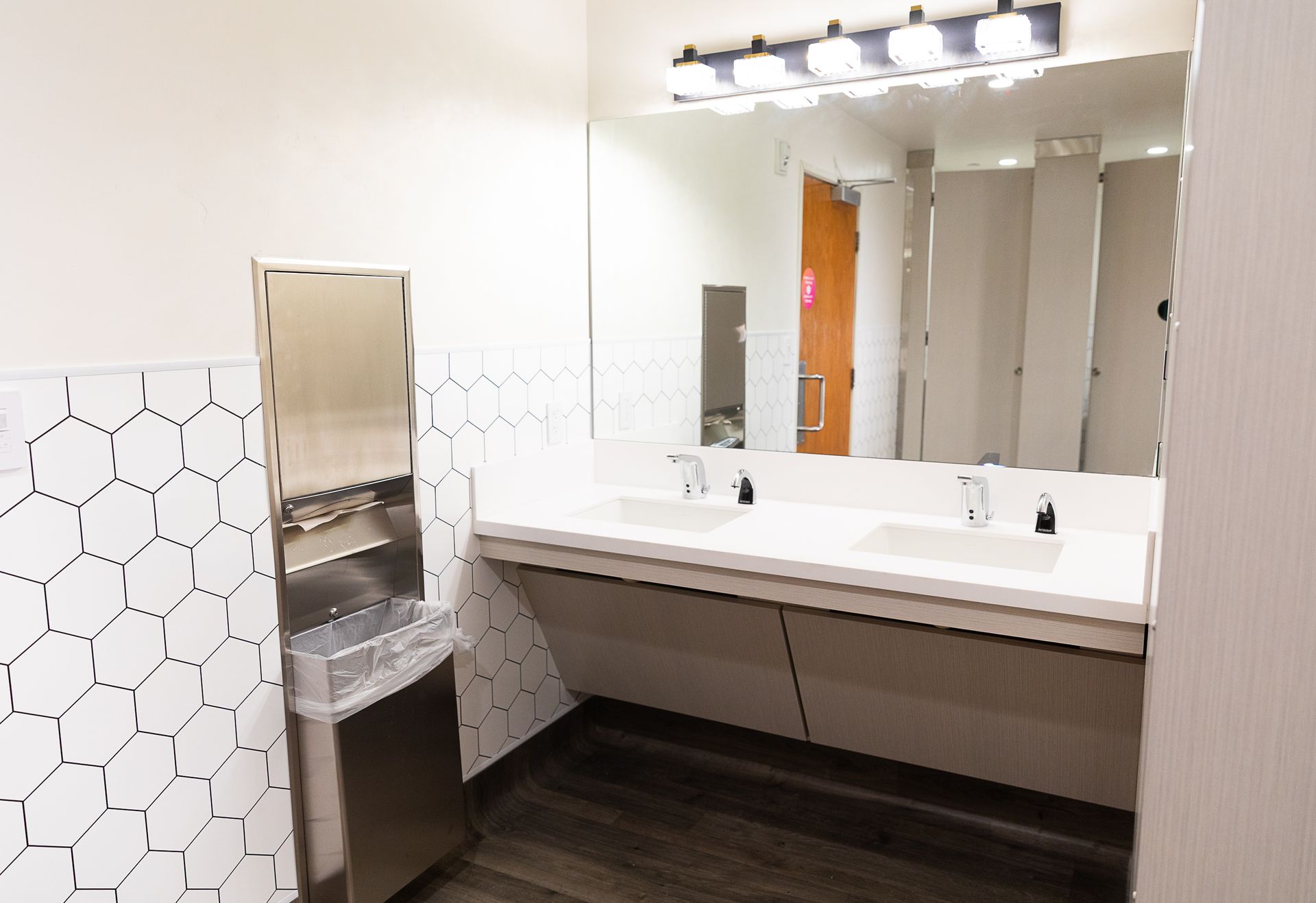 Accessible restroom with white hex tile wall, long mirror, sinks, and a metal drinking fountain or hand dryer