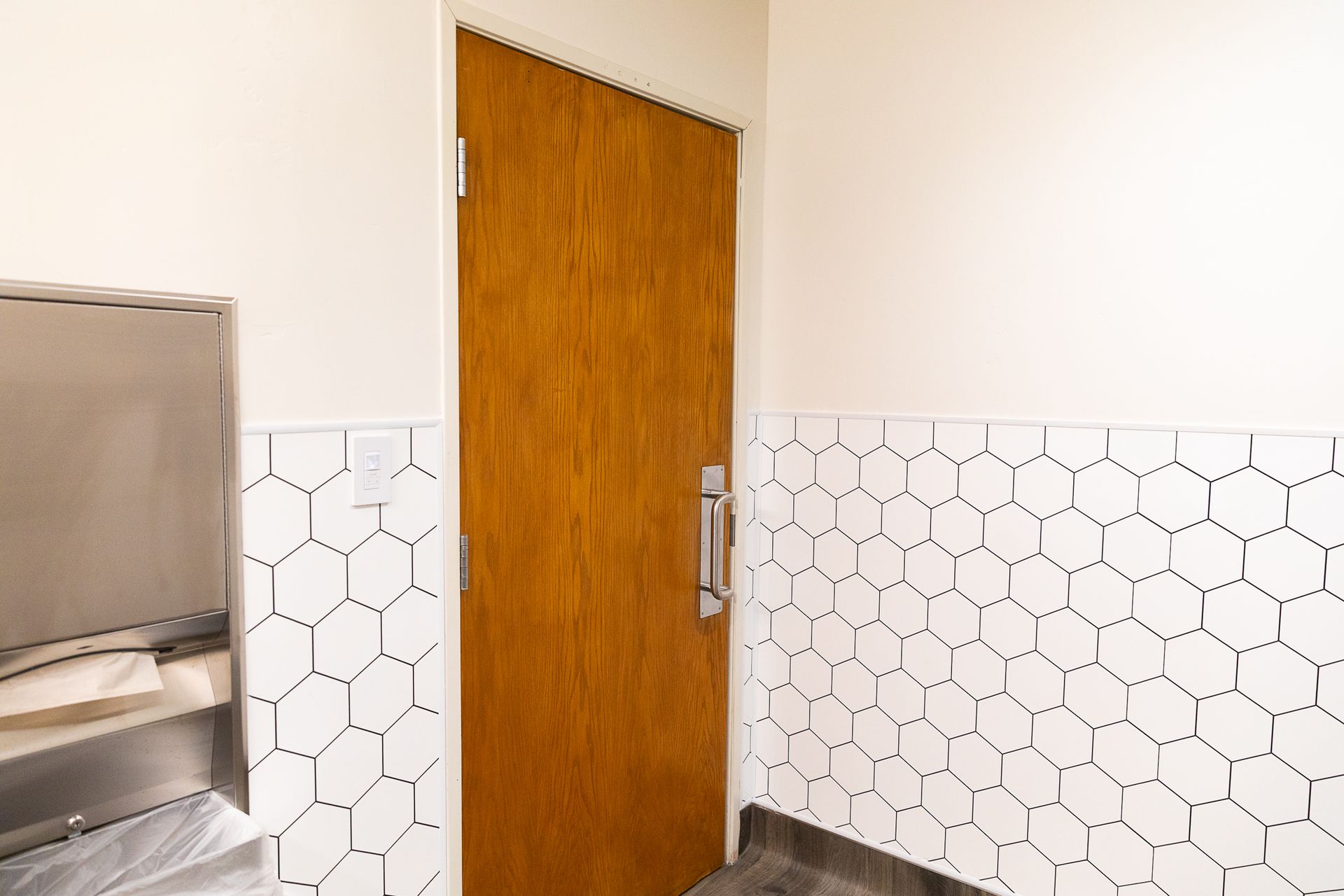 White-tiled hallway with a closed wooden door and a stainless steel dispenser on the left.