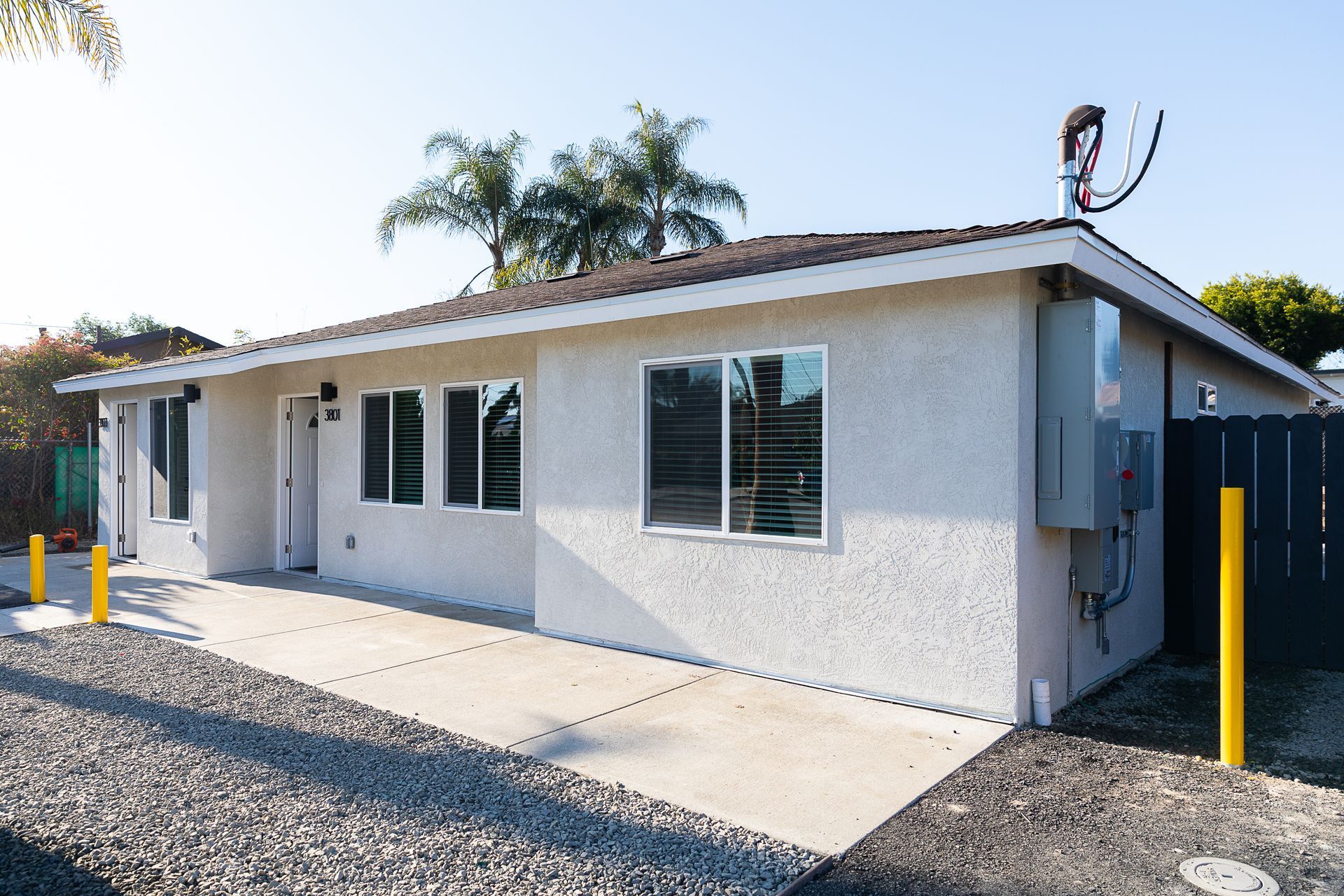 Single-story white stucco house with windows, gravel driveway, and yellow bollards in bright sunlight