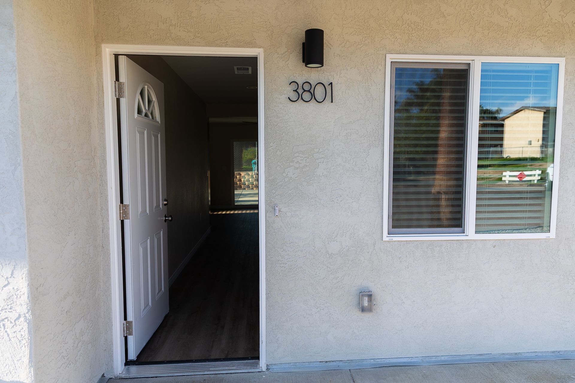 Front entrance of a stucco house with open door, window, porch light, and house number 3801