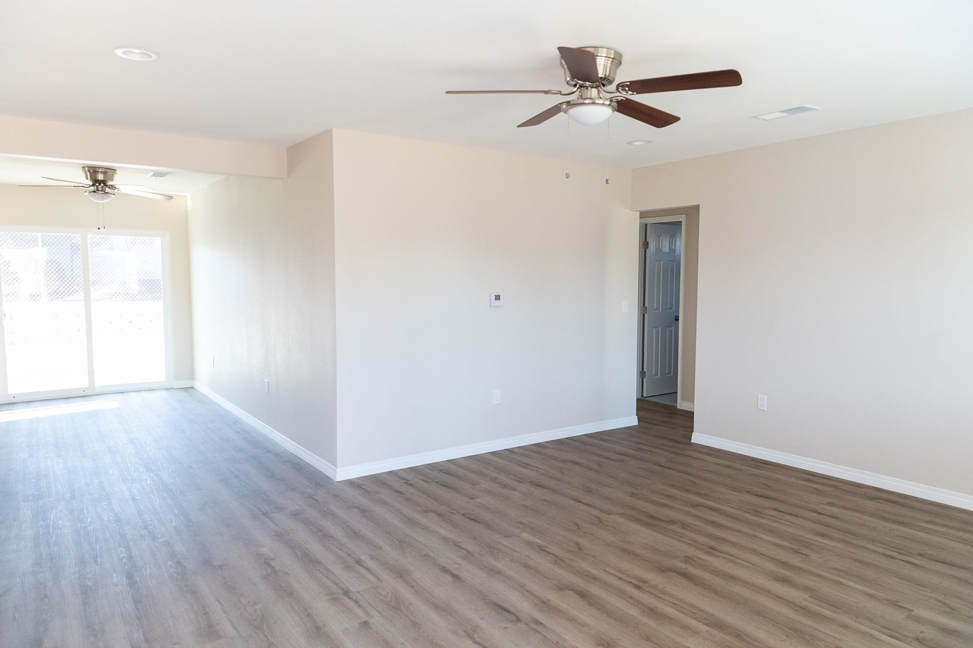 Empty living room with wood-look floors, white walls, ceiling fan, and sliding glass doors to a patio