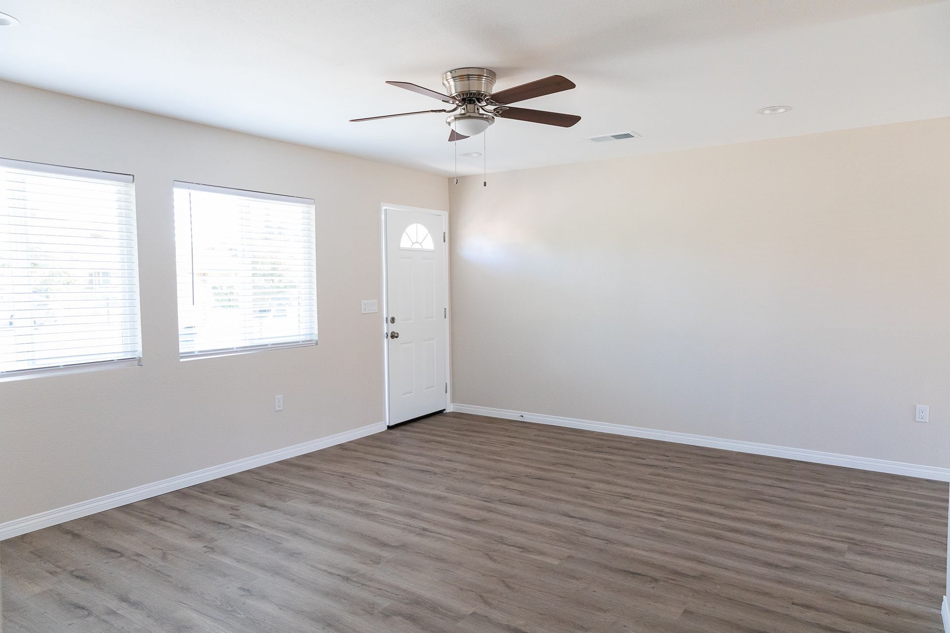 Empty living room with white walls, wood-look flooring, windows, a ceiling fan, and a front door