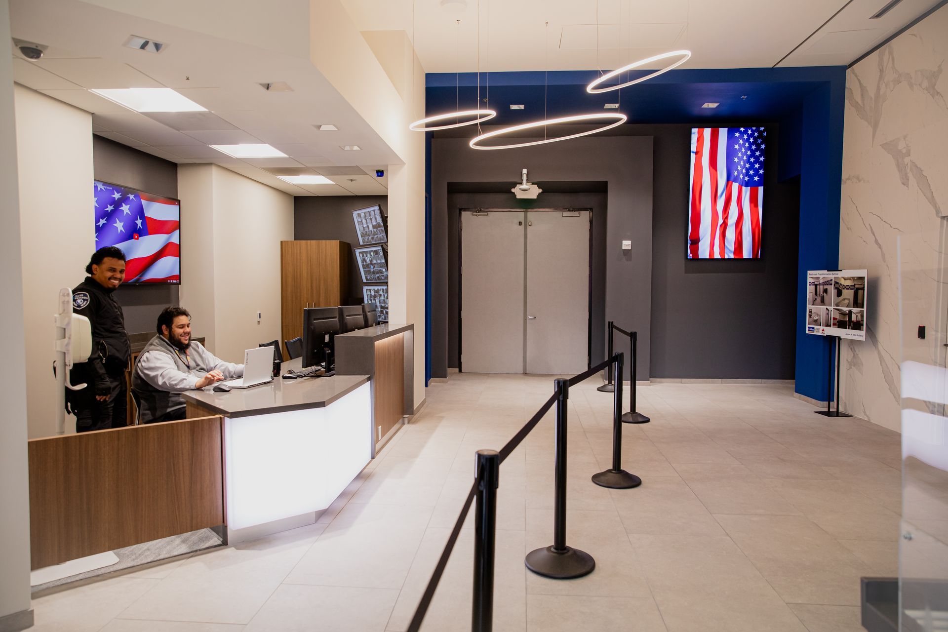 Reception desk in a modern lobby with two staff members and American flags on wall screens.