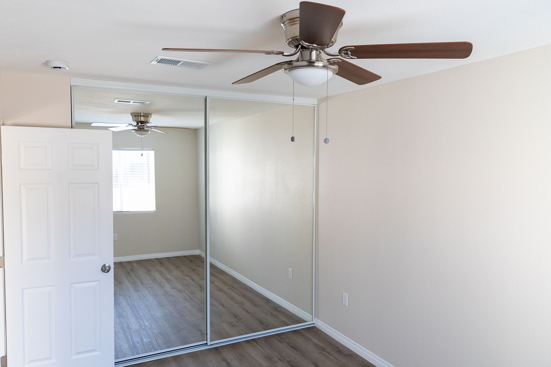 Empty bedroom with mirrored closet doors, white walls, wood floor, and a ceiling fan