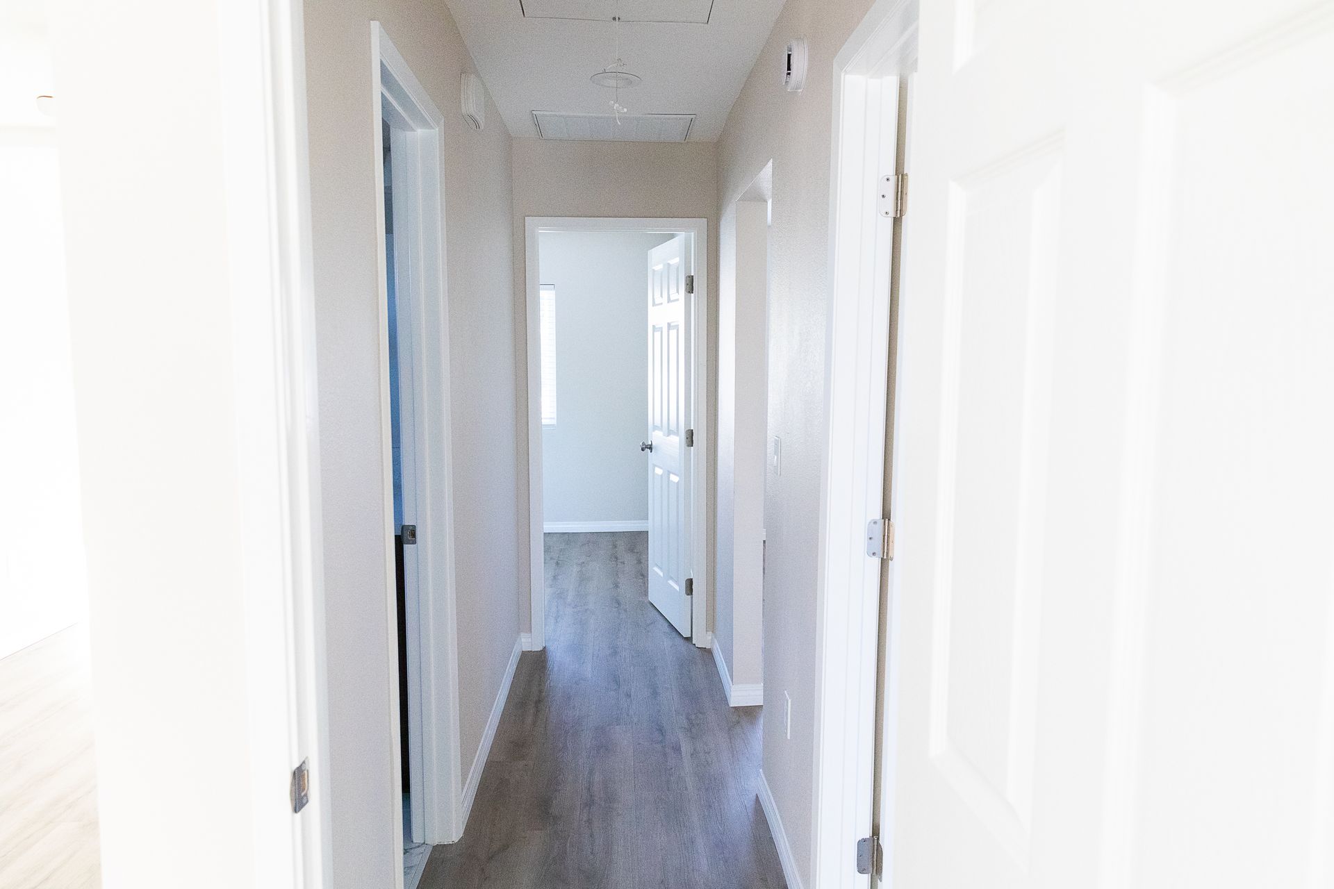 Bright white hallway with gray flooring and doors on both sides, leading to an open doorway.