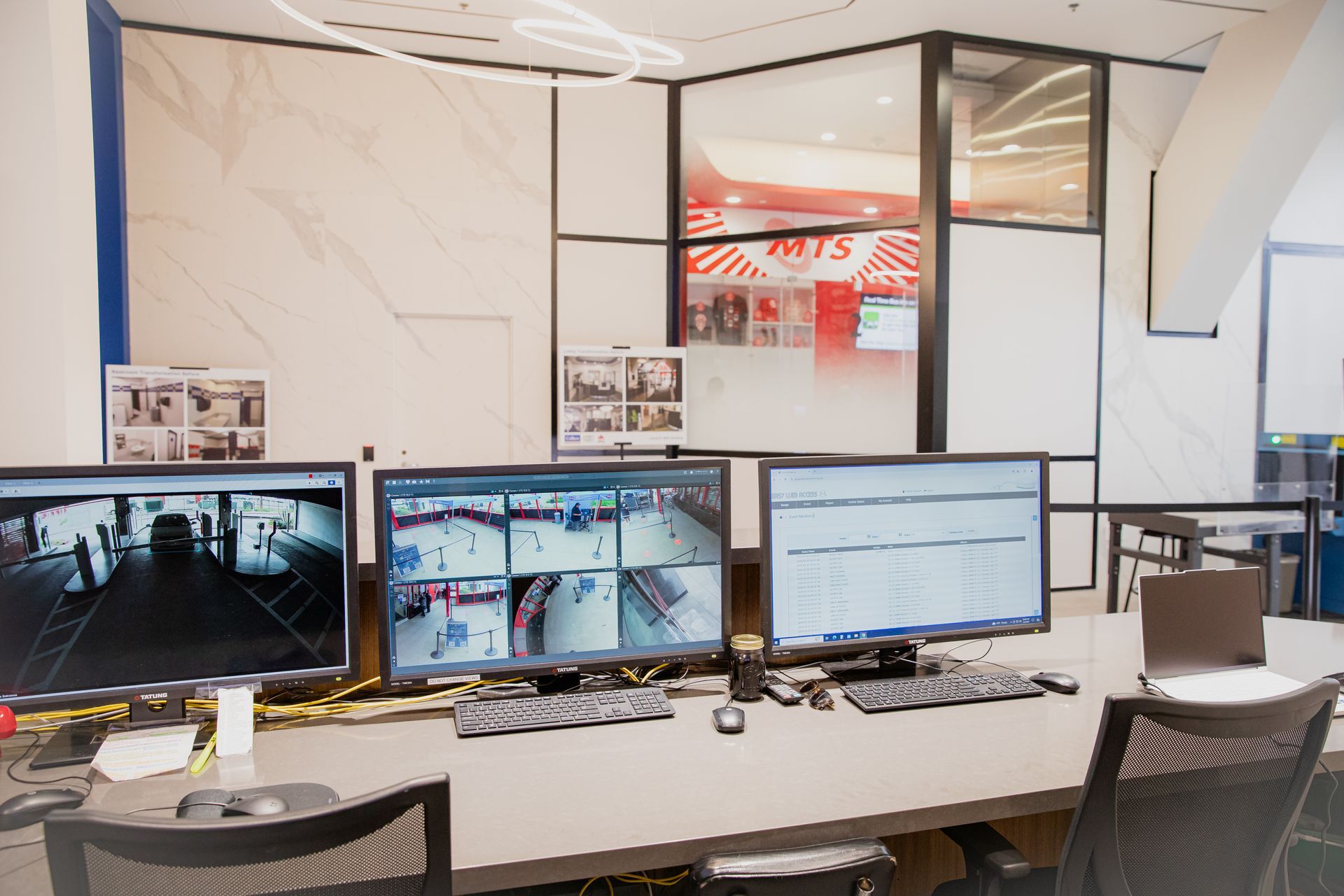Modern office control room with four monitors on a desk and empty chairs, overlooking a bright interior space