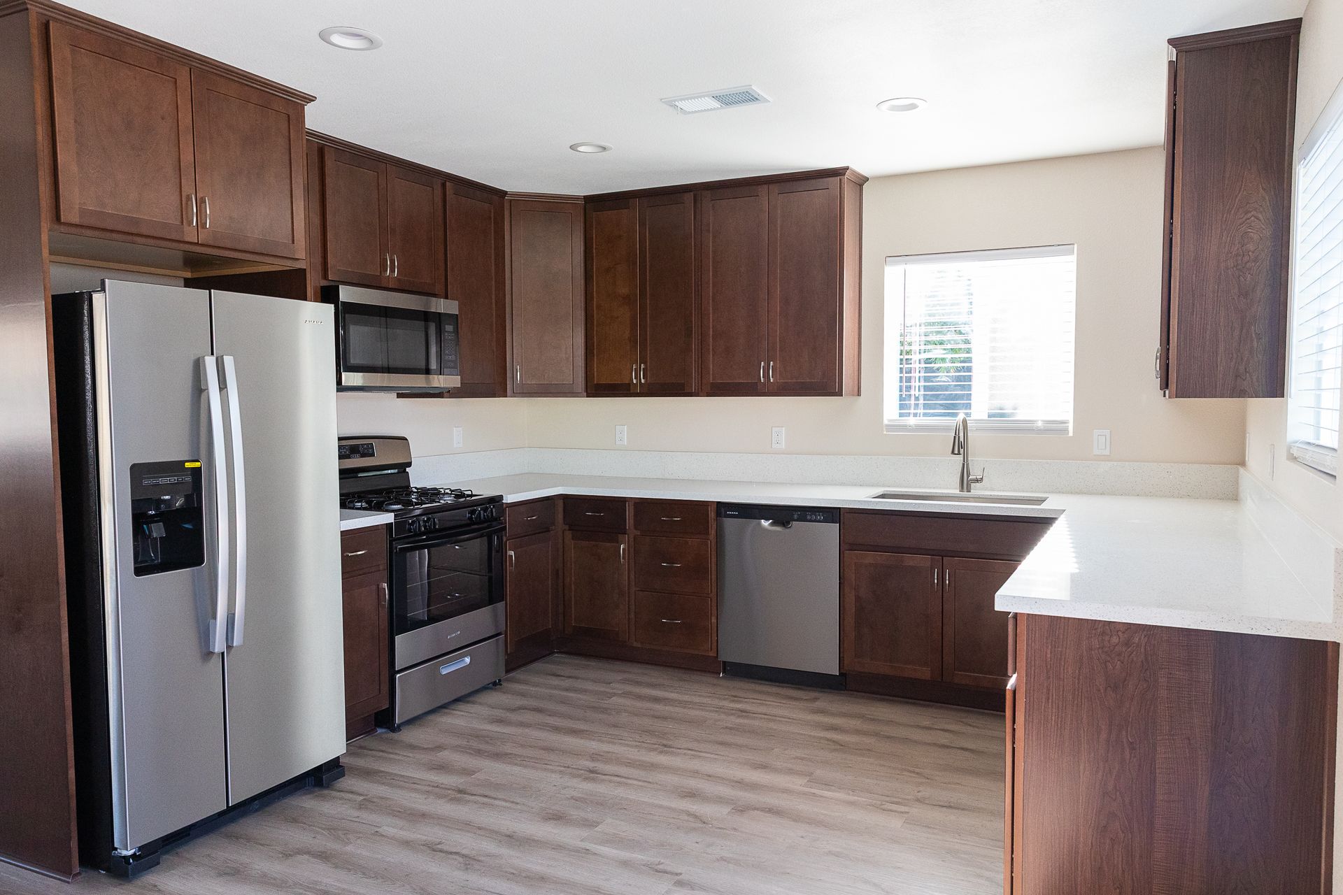 Empty kitchen with dark wood cabinets, stainless steel refrigerator, white countertops, and a window over the sink