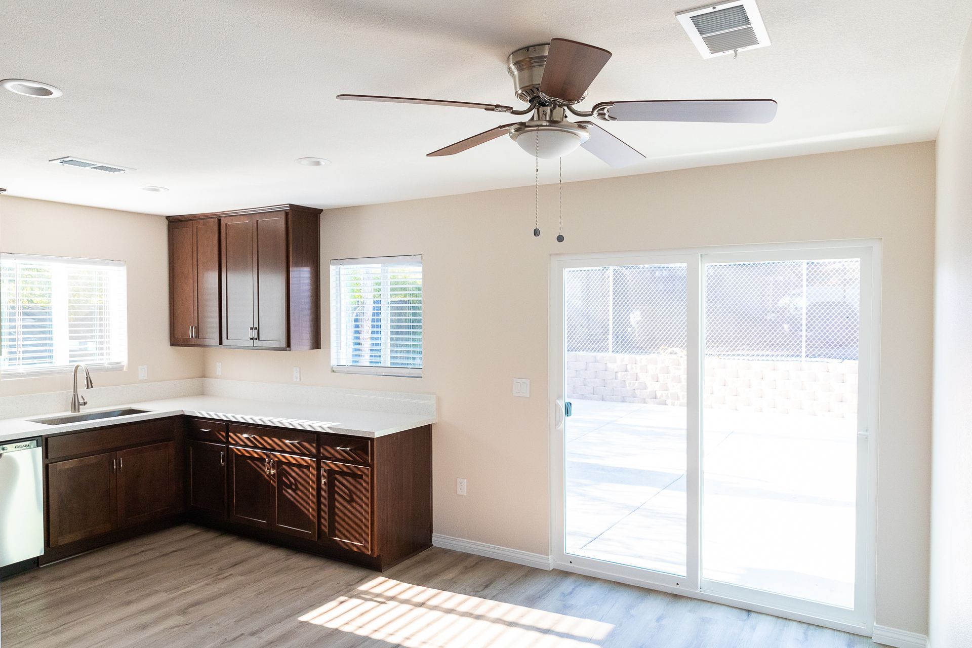 Bright empty kitchen with dark wood cabinets, white counters, ceiling fan, and sliding glass doors to a patio.