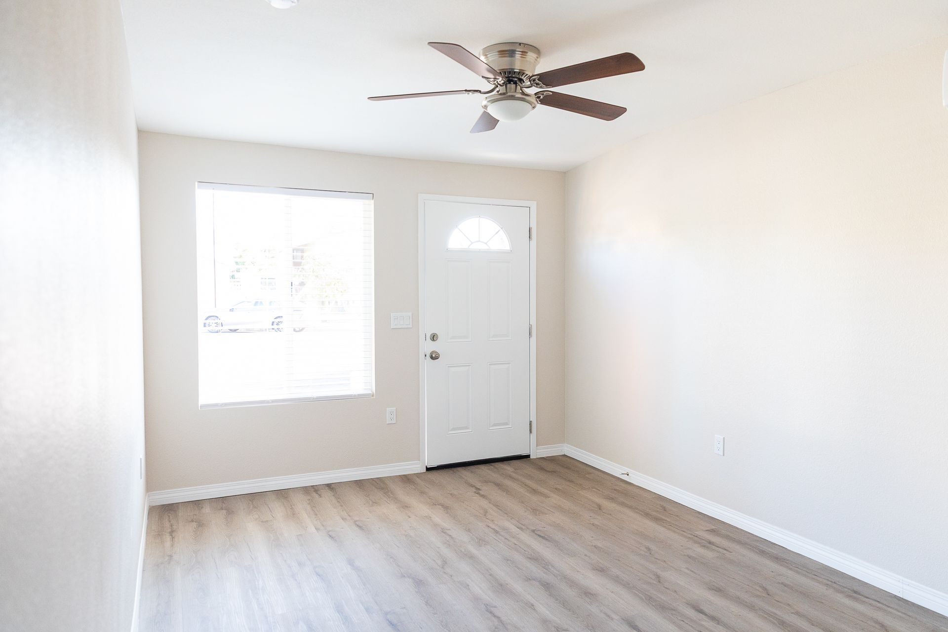 Empty bright room with white walls, wood-look floor, window, front door, and ceiling fan.