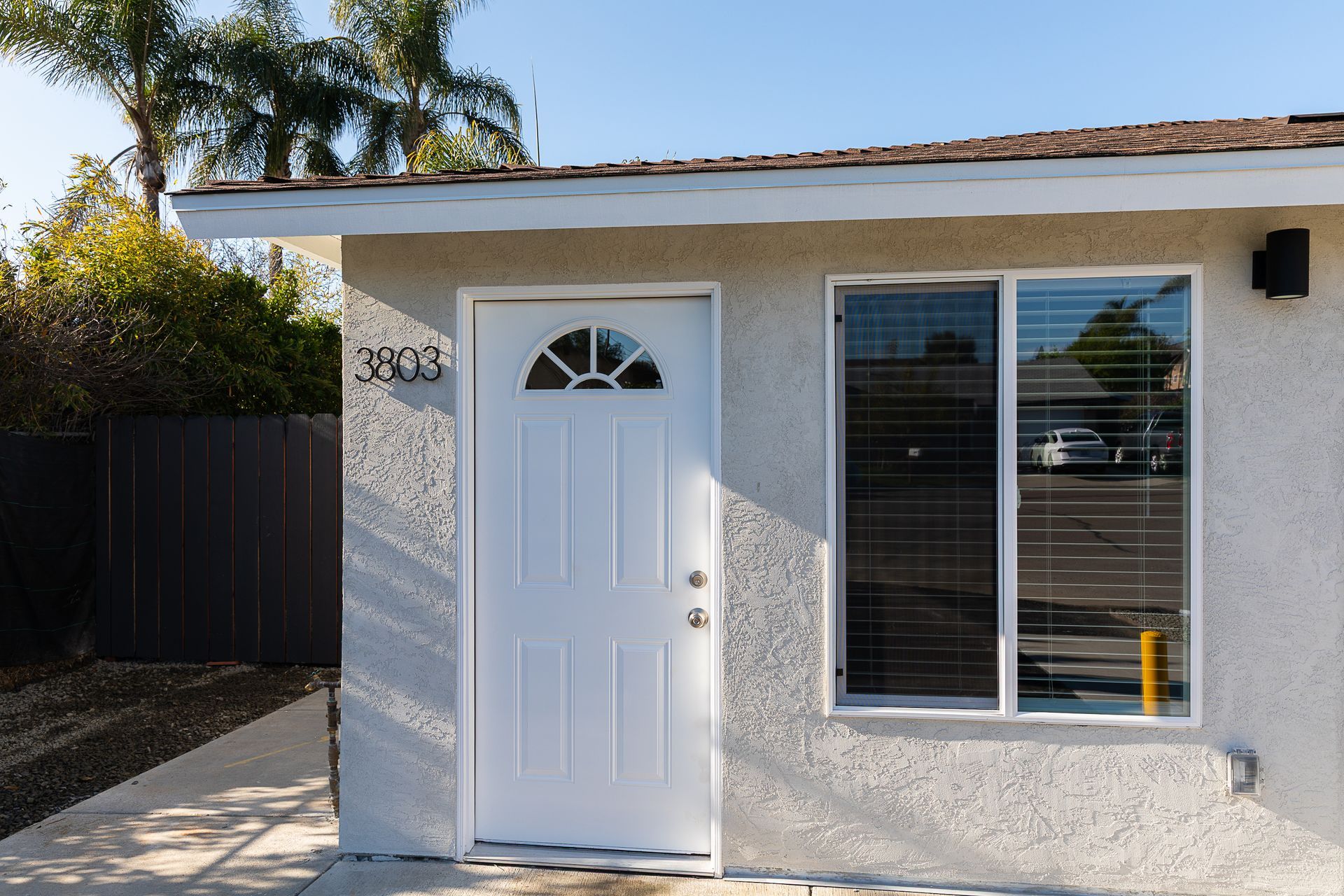Small white stucco shed with a white door and window, beside a black fence in a sunny yard