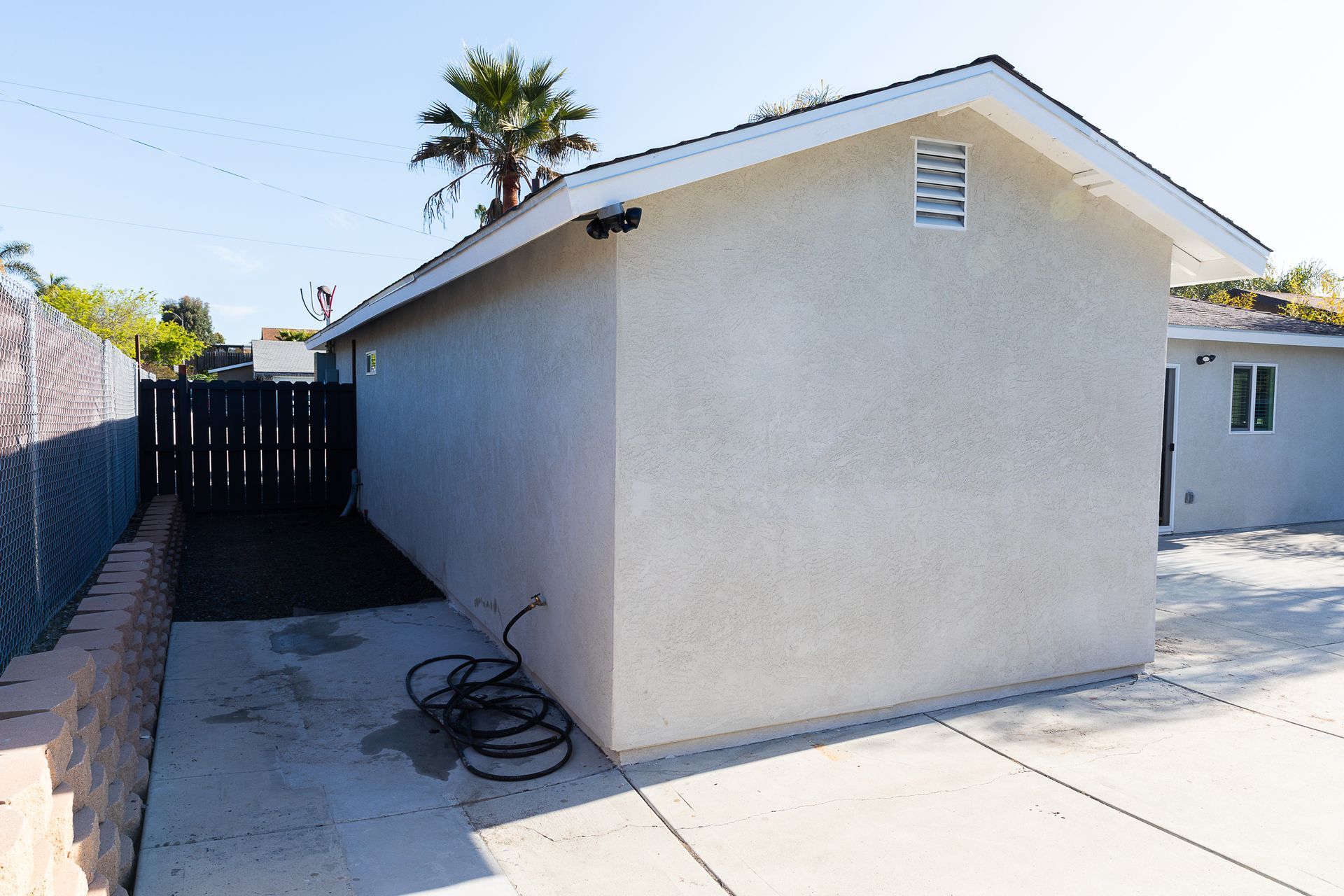 Side yard beside a beige house, with concrete driveway, black gate, and coiled hose on the ground