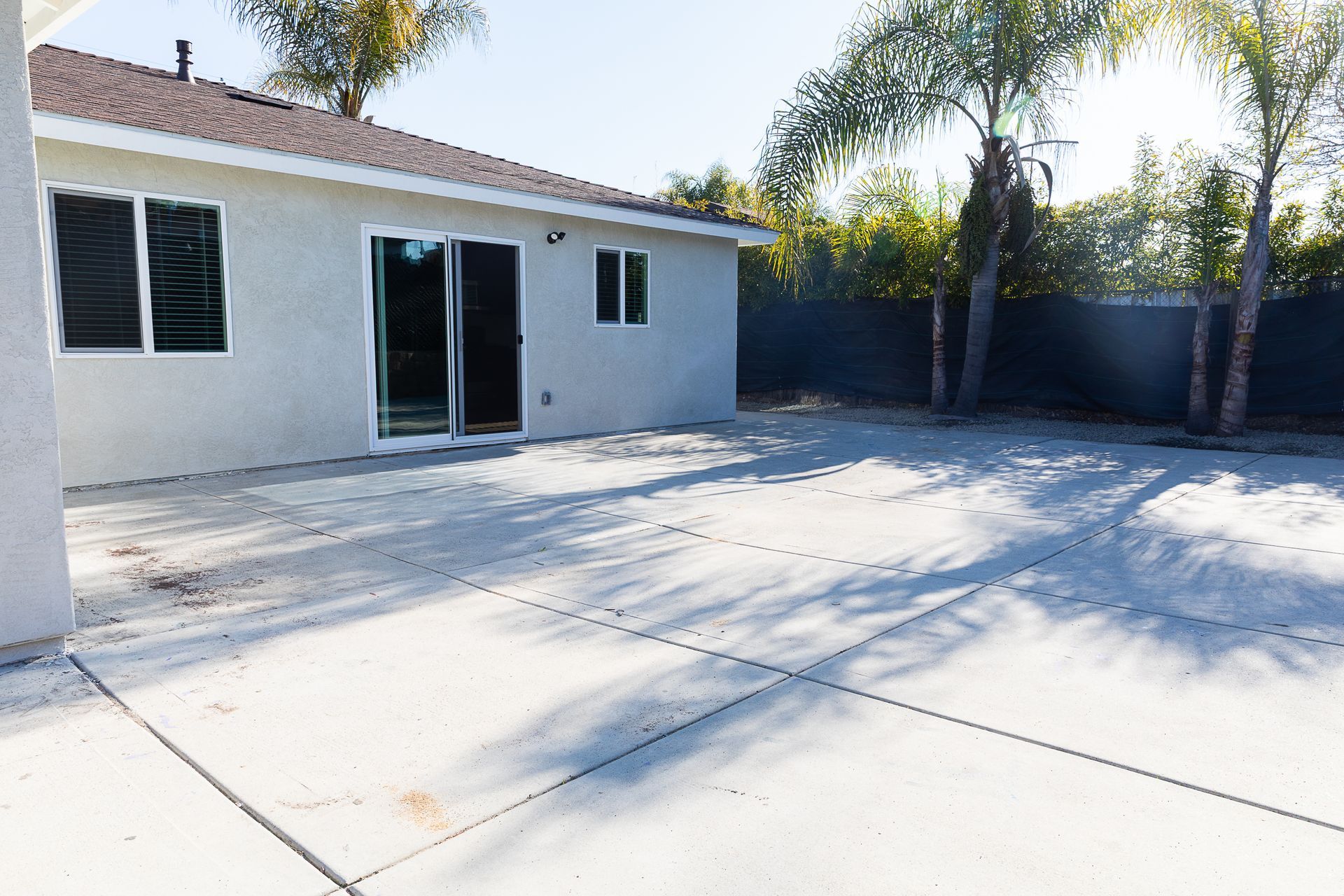 Backyard patio with a white house, sliding glass door, and concrete slab under bright sunlight