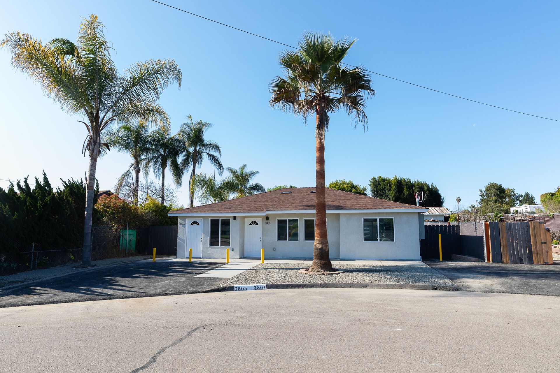 Single-story white house with a red-tiled roof, palm trees, and a gravel driveway under a clear blue sky