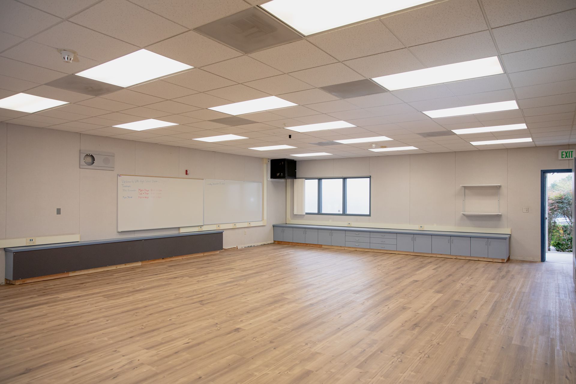 Empty classroom with wood floor, whiteboard, cabinets, and fluorescent ceiling lights