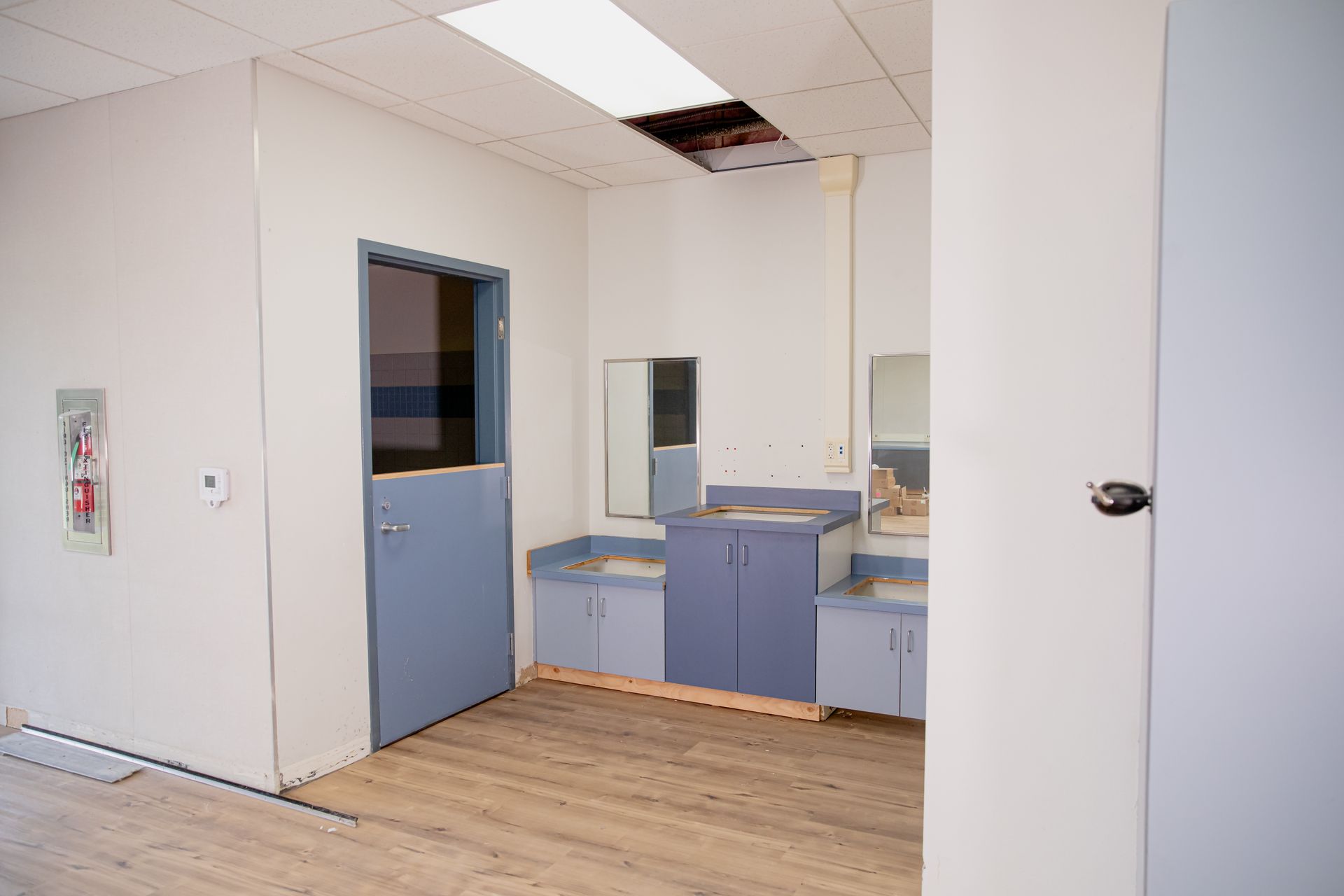 Empty office kitchenette with blue cabinets, white walls, and a damaged drop ceiling panel
