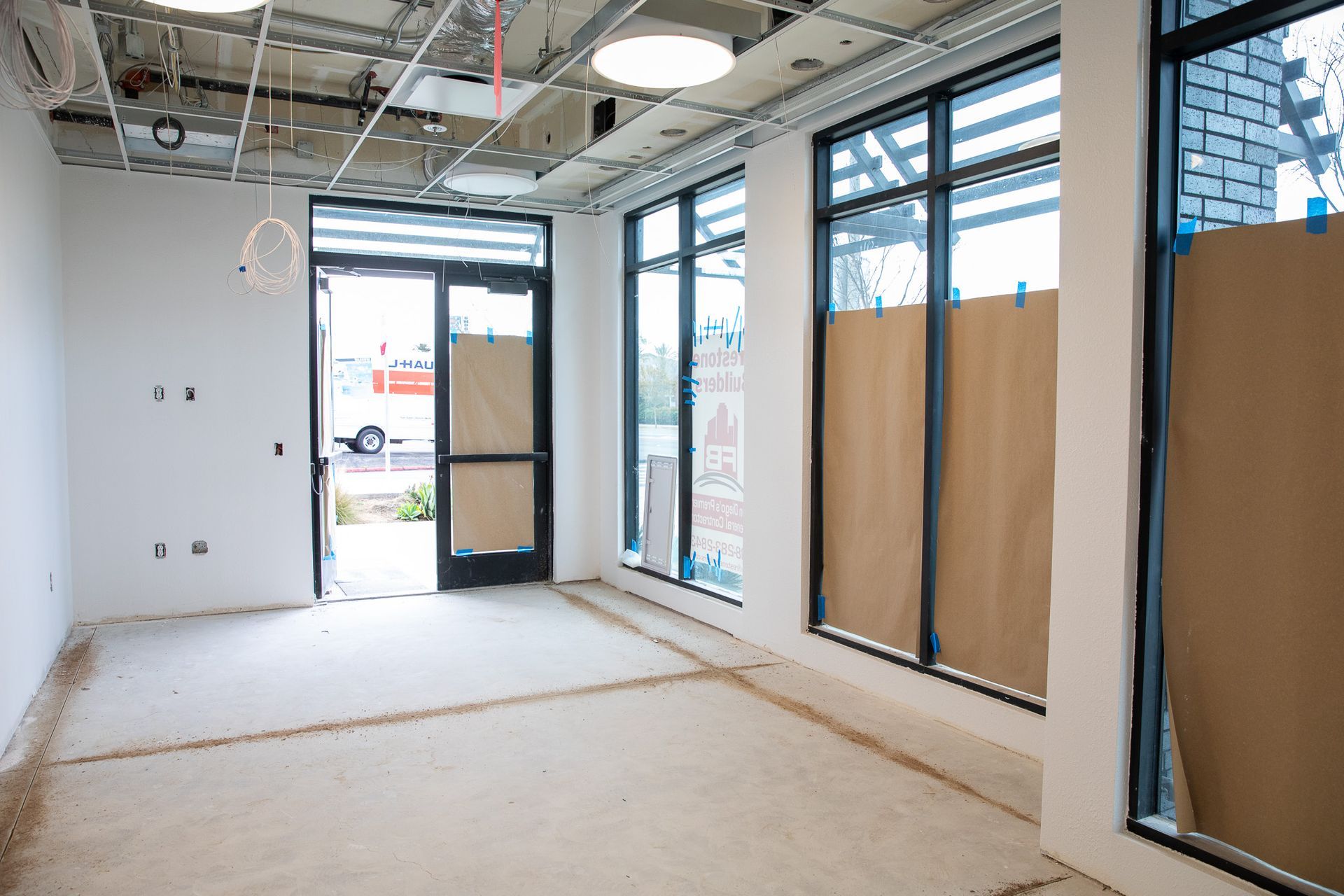 Empty unfinished storefront interior with exposed ceiling, large windows, and boarded door and glass panels