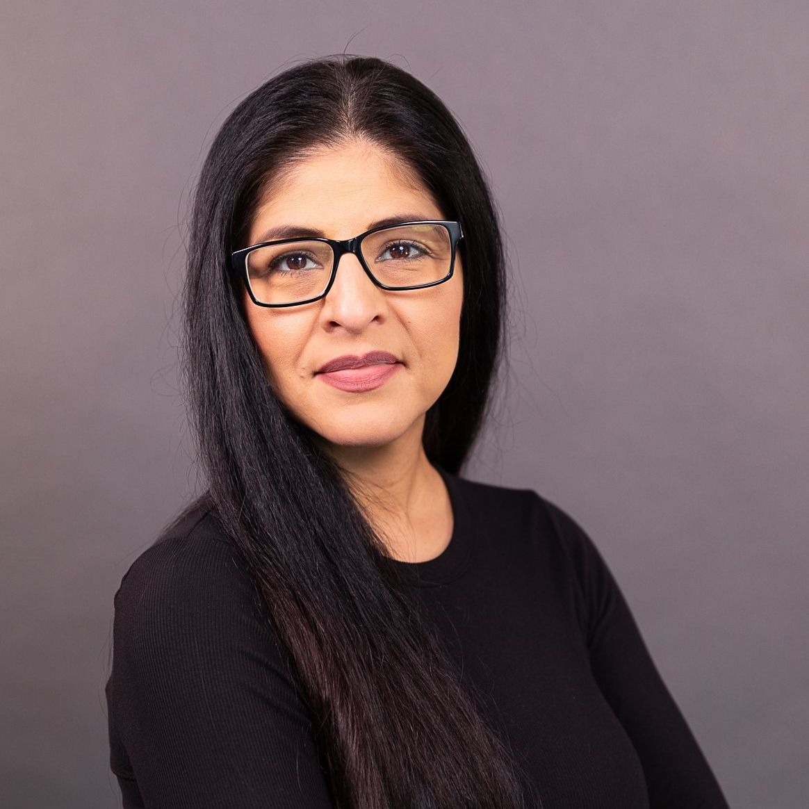 Portrait of a person with long dark hair and glasses, wearing a black top against a gray background