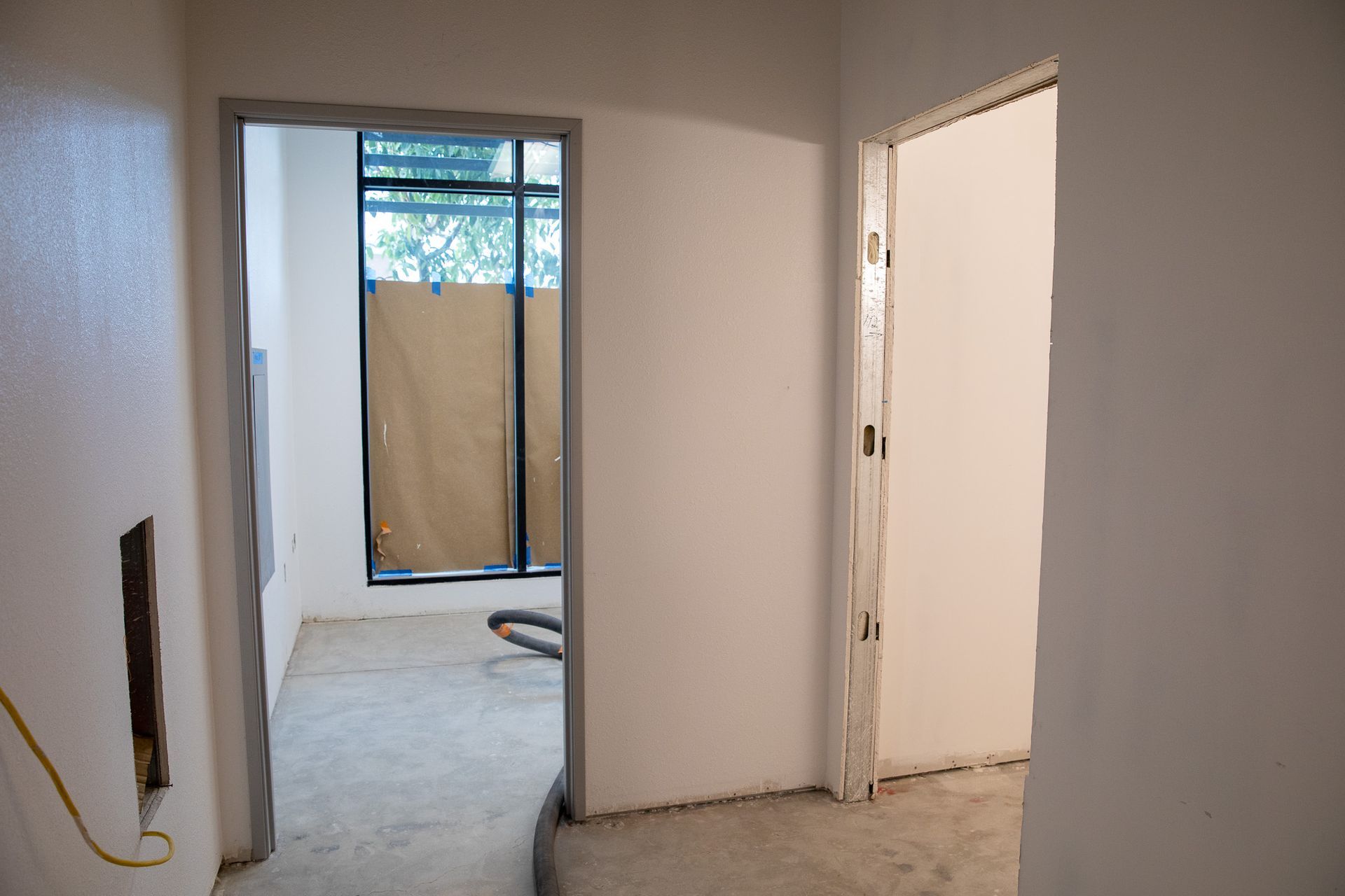 Empty unfinished hallway with white walls, doorways, and a windowed opening to a tiled outdoor area.
