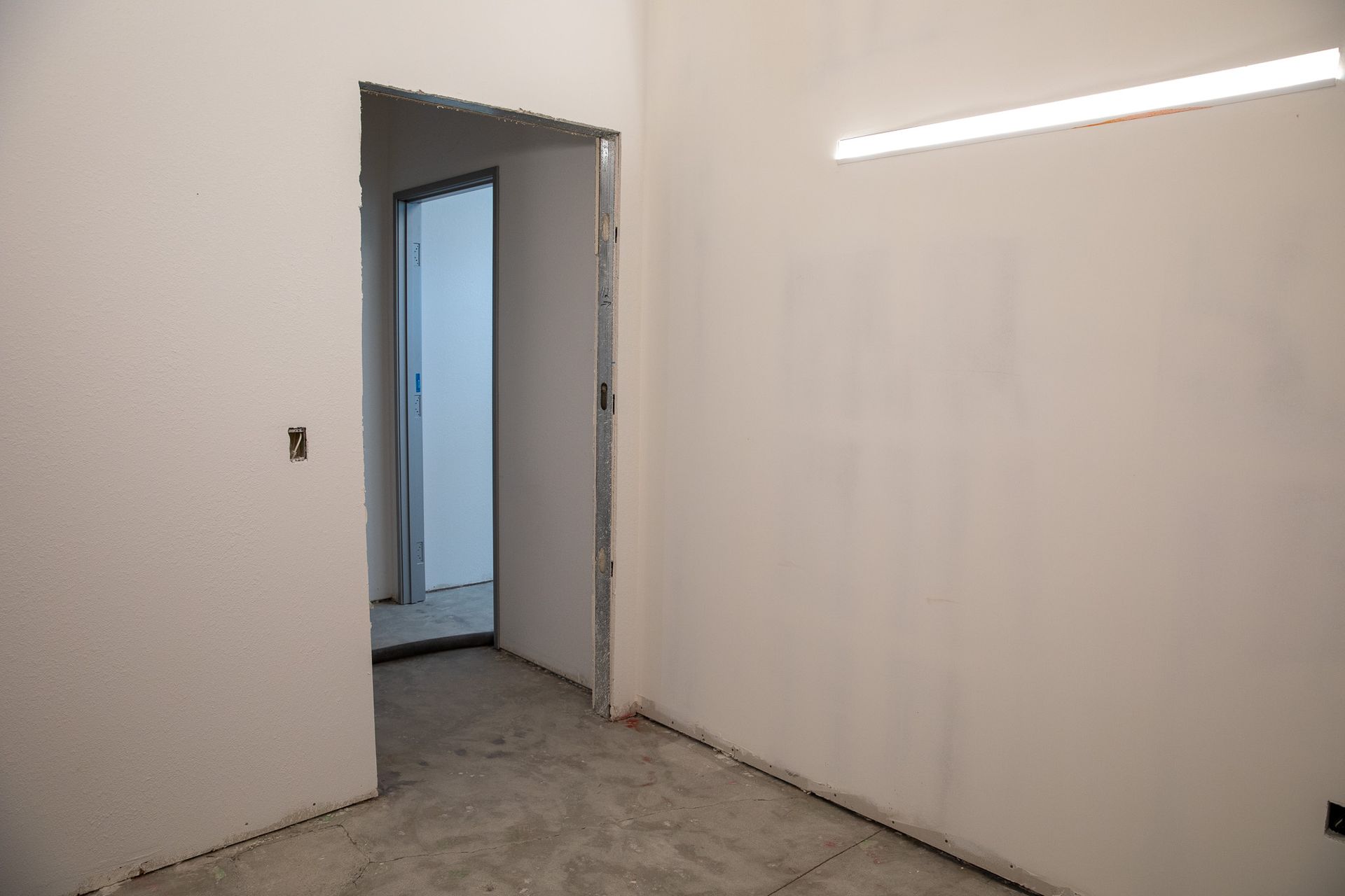 Empty unfinished room with white walls, gray floor, and open doorway to a blue-lit hall