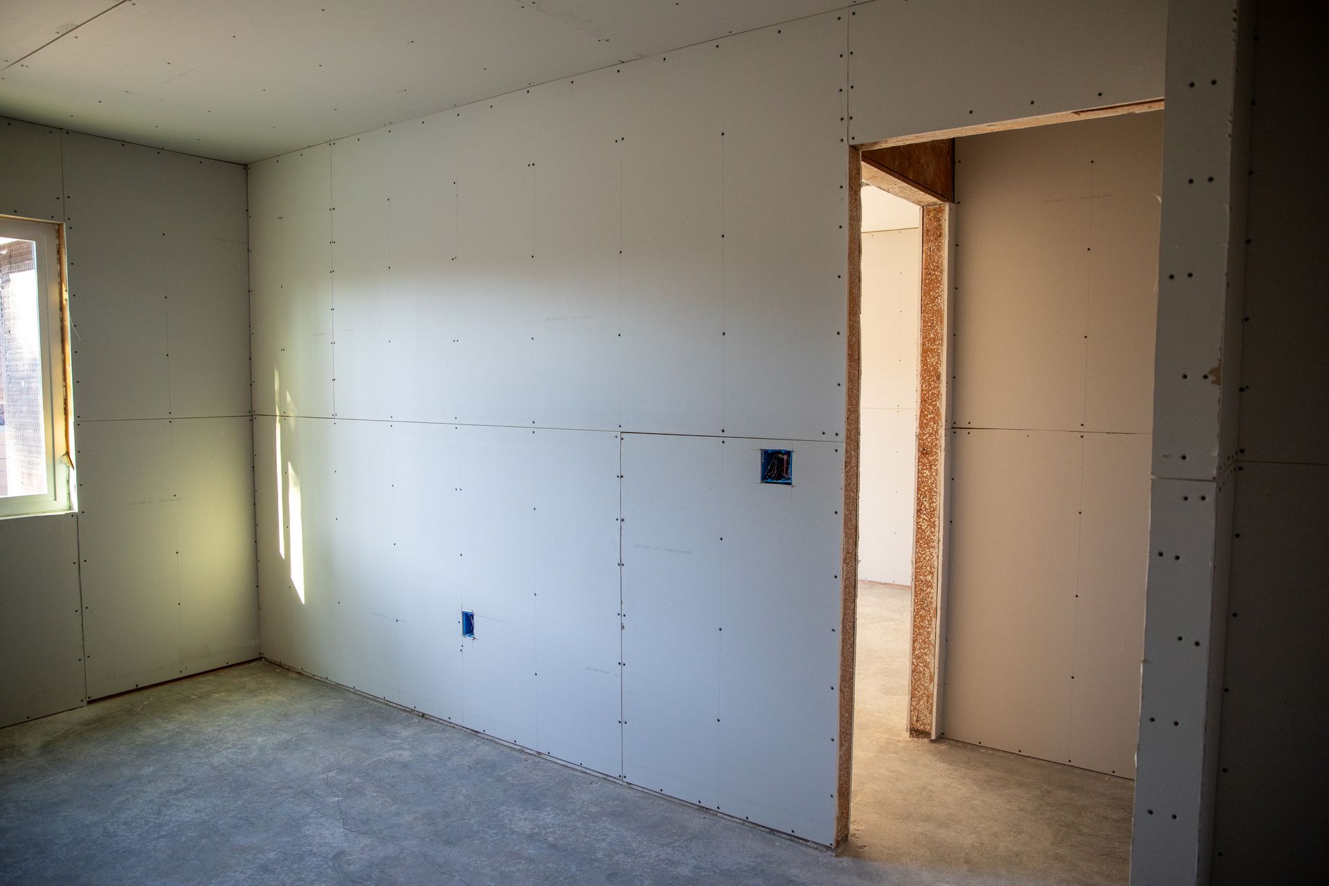 Unfinished room with drywall, a window on the left, and an open doorway to another room.