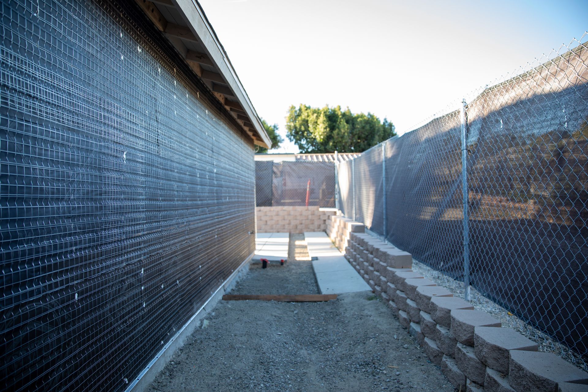Narrow gravel walkway between black mesh fences, with a concrete step and trees in the distance.