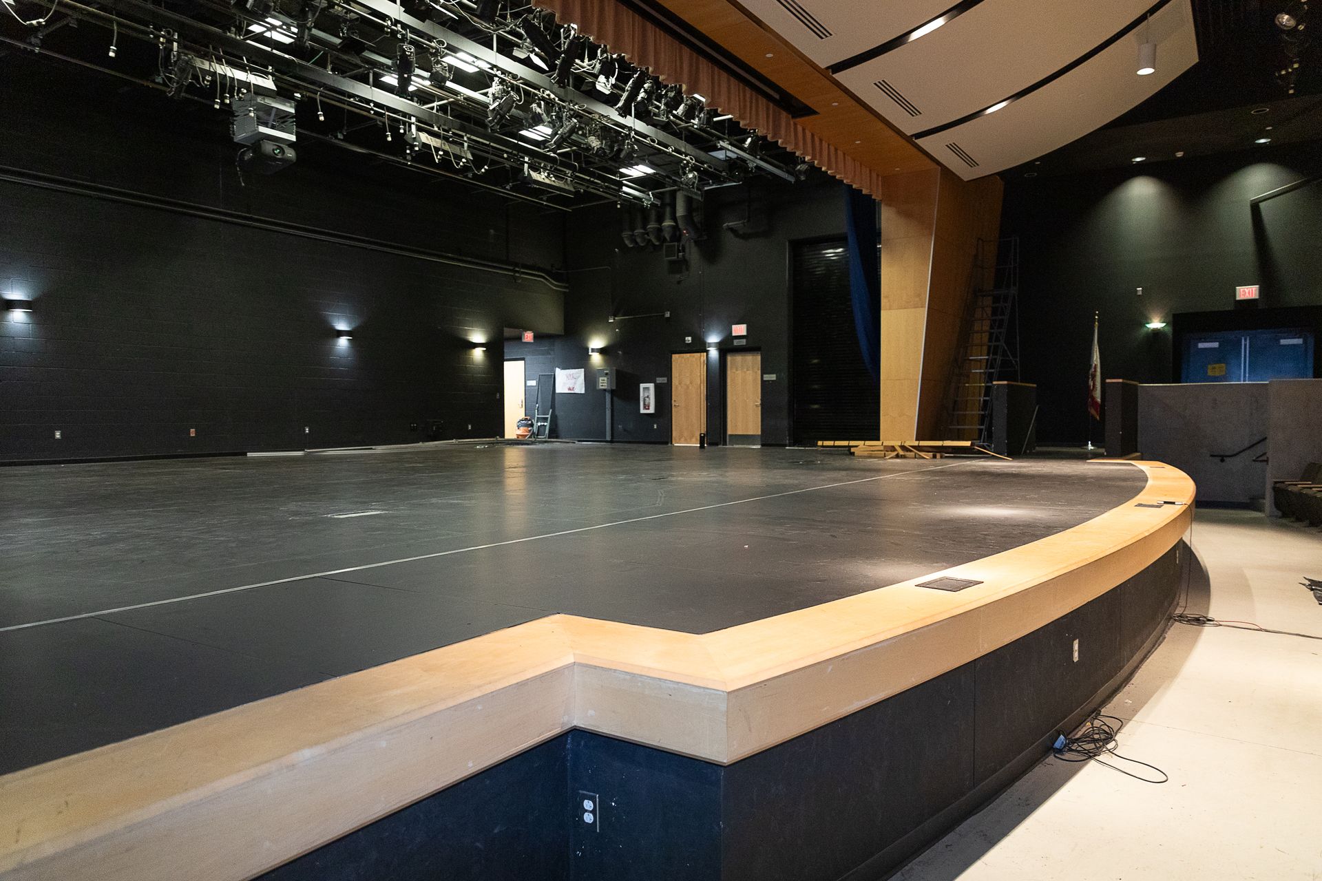 Empty indoor stage with black backdrop, overhead lights, and curved front edge