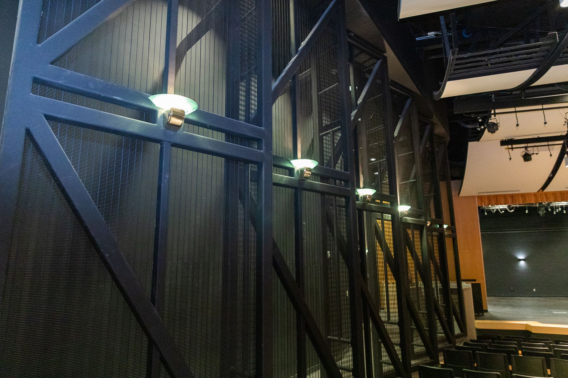 Dim indoor stage with black metal mesh walls, hanging lights, and rows of seating.