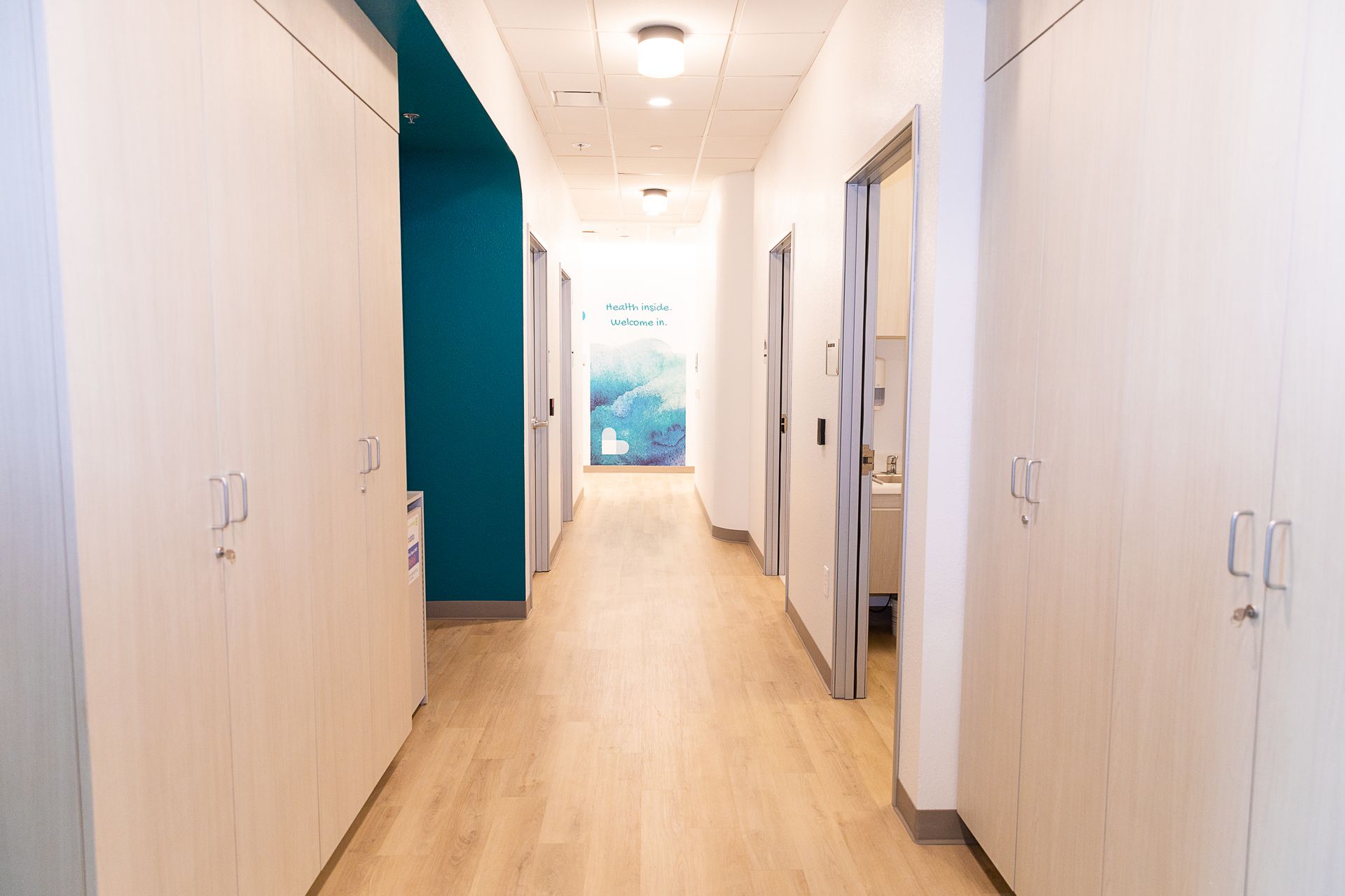 Bright hallway with wood floors, white cabinets, and a teal accent wall leading to a windowed doorway