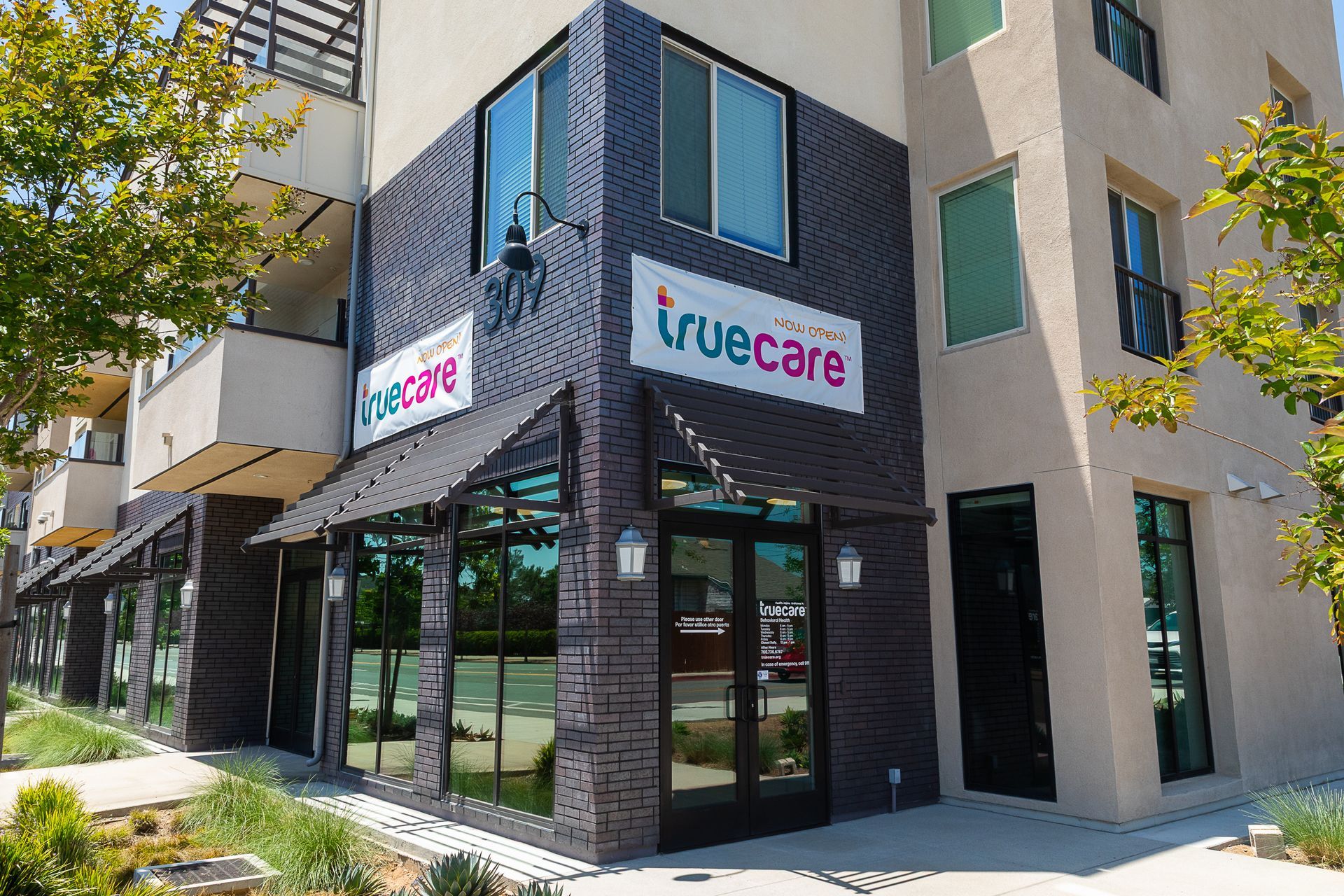 Corner storefront of TrueCare with dark brick facade and glass windows on a sunny street.