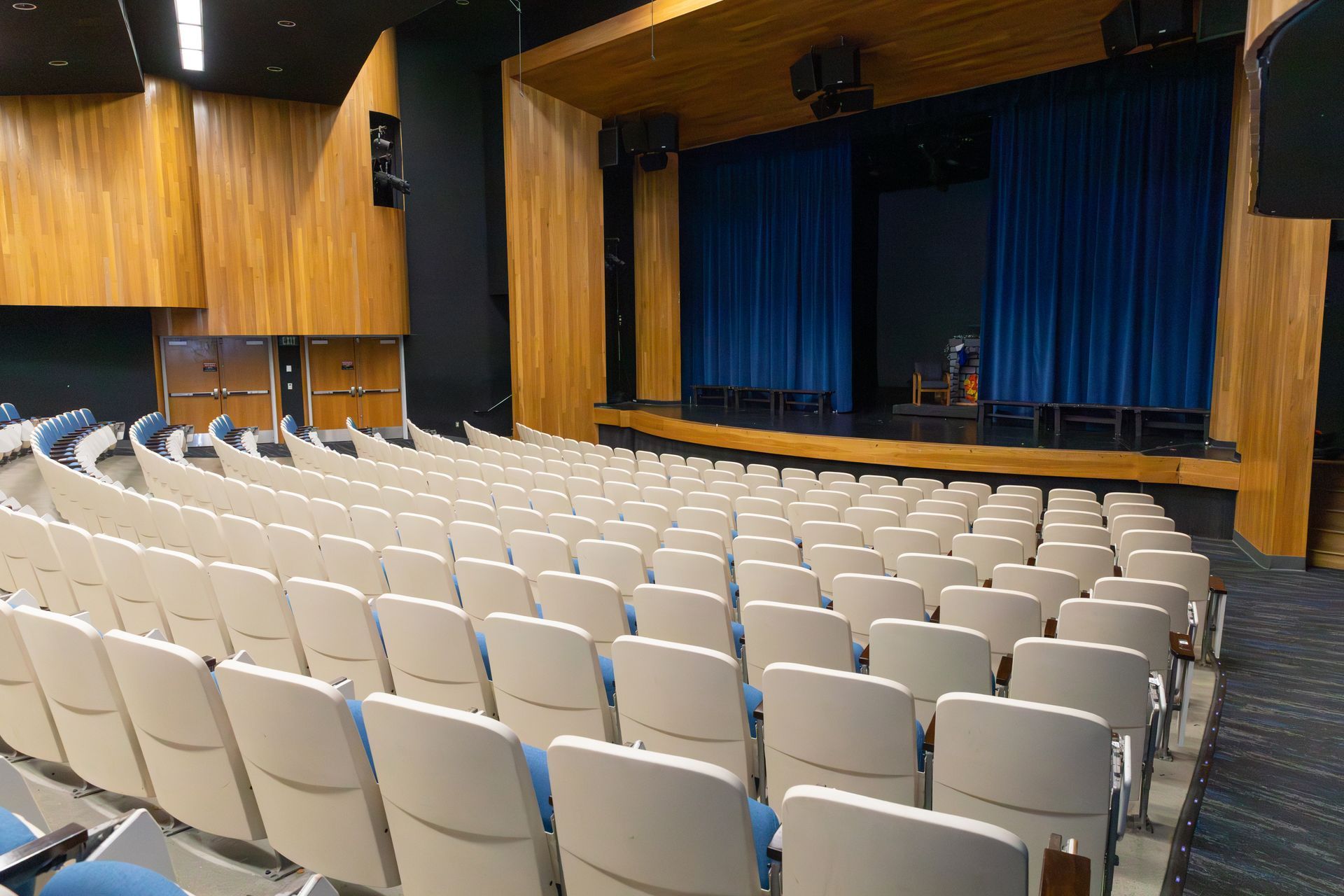 Empty auditorium with rows of beige seats facing a stage with blue curtains and wood-paneled walls