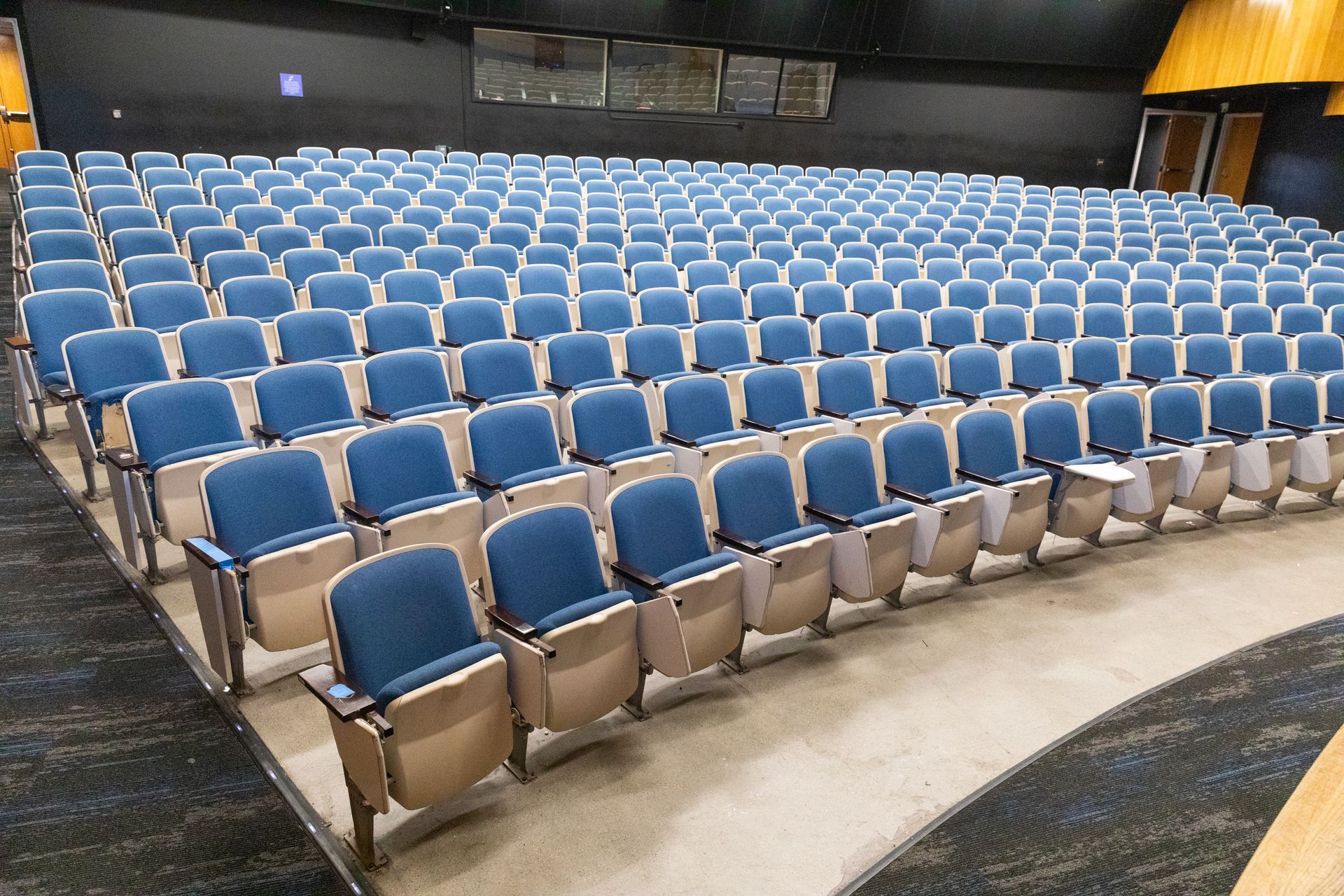 Empty auditorium with rows of blue seats and tan armrests facing a dark stage