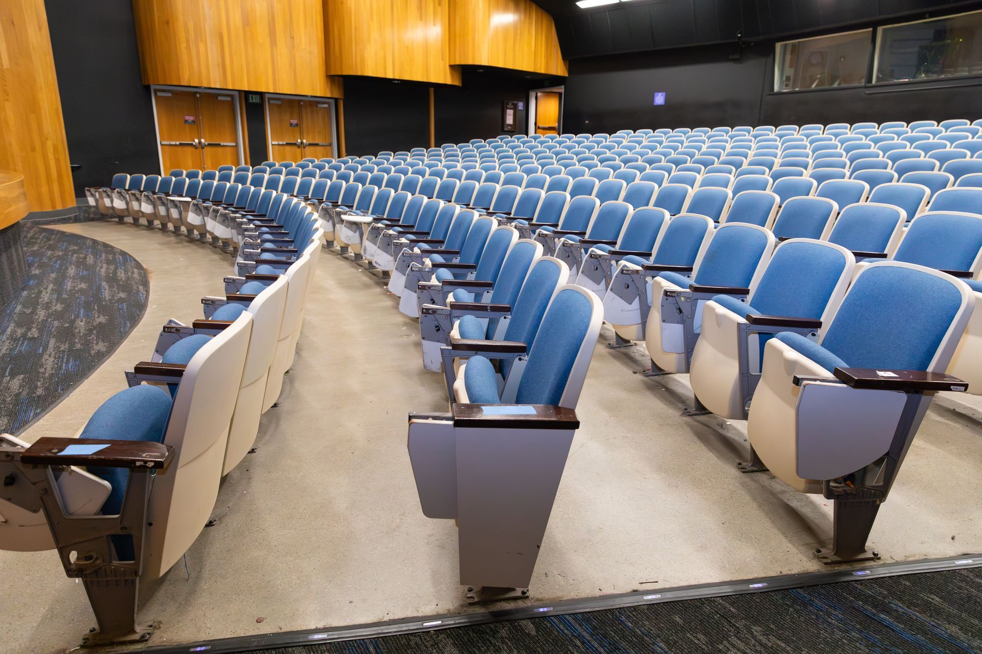 Empty curved lecture hall with rows of blue seats and a wood-paneled stage area