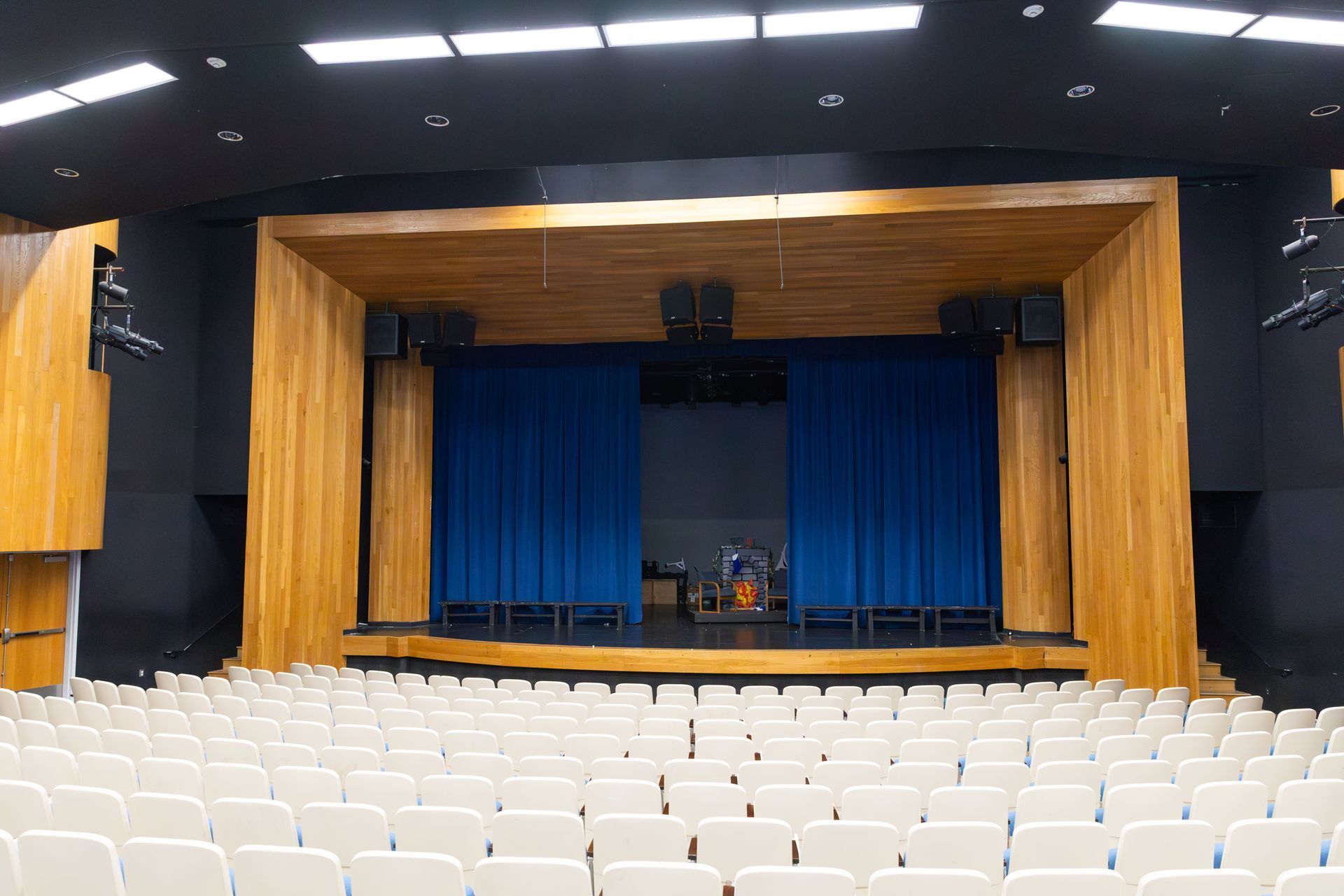 Empty auditorium with rows of white seats facing a stage with blue curtains and wood paneling