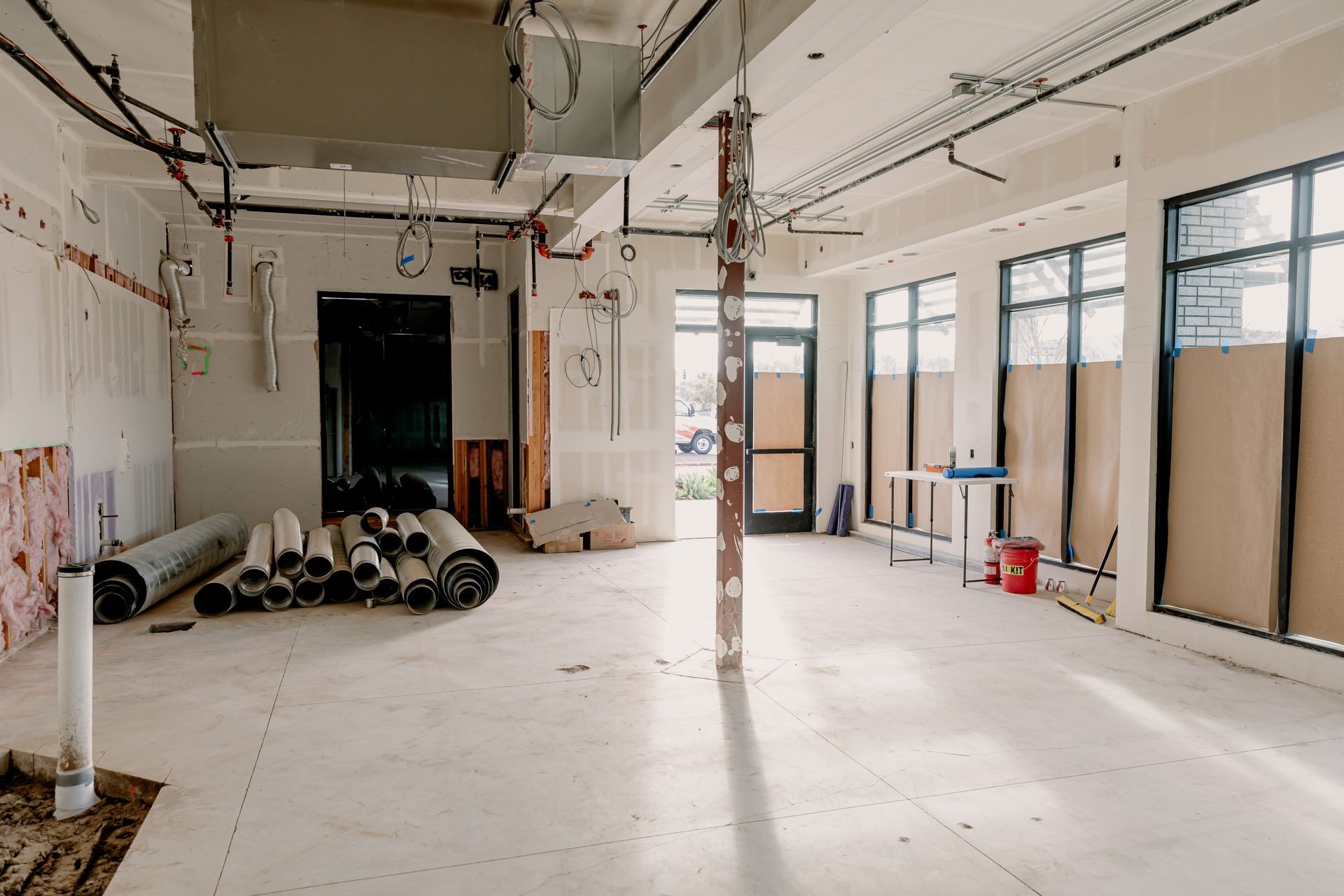 Empty unfinished room with white walls, exposed ceiling, pipes, and construction materials on the floor.