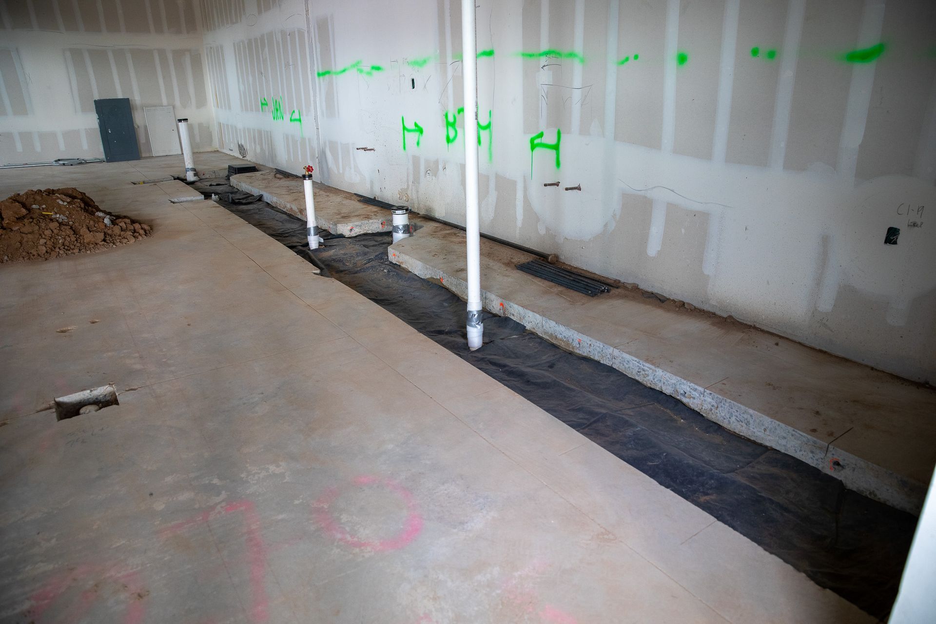 Interior construction site with drywall, exposed pipes, and a long floor trench along the wall